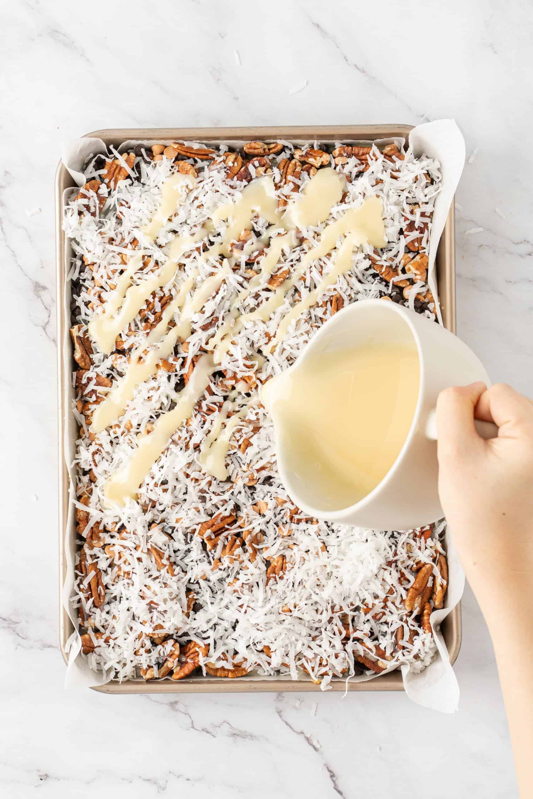 Sweetened condensed milk being poured over coconut, pecans, and chocolate chips for magic bars.