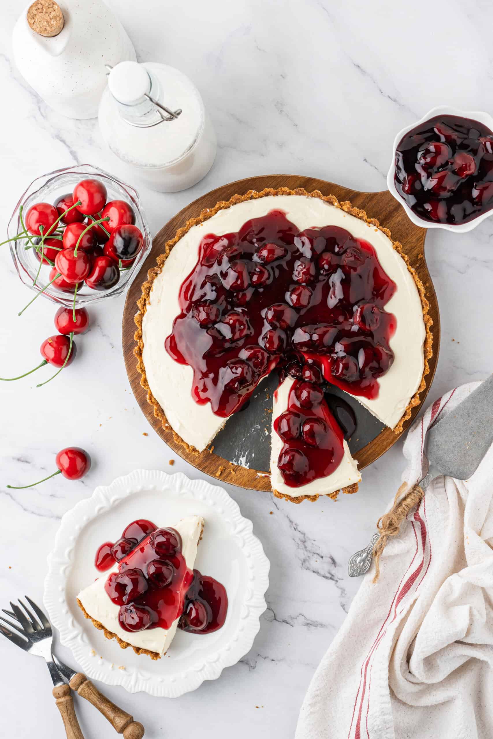 Overhead view of a no-bake cheesecake topped with cherry pie filling, with one slice removed and another served on a dessert plate.
