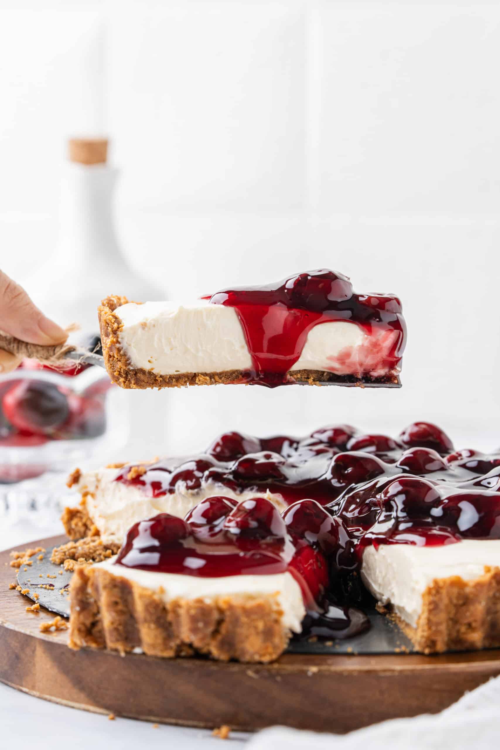 Slice of no-bake cheesecake with graham cracker crust and cherry pie filling being lifted from the cheesecake on a cake server.