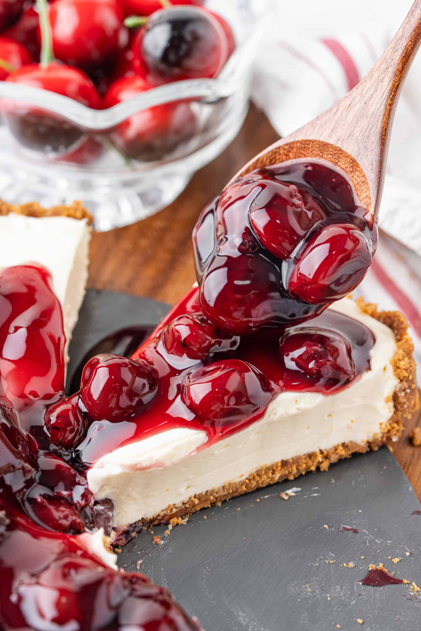 Spoonful of cherry pie filling being added to a slice of no-bake cheesecake with a graham cracker crust.