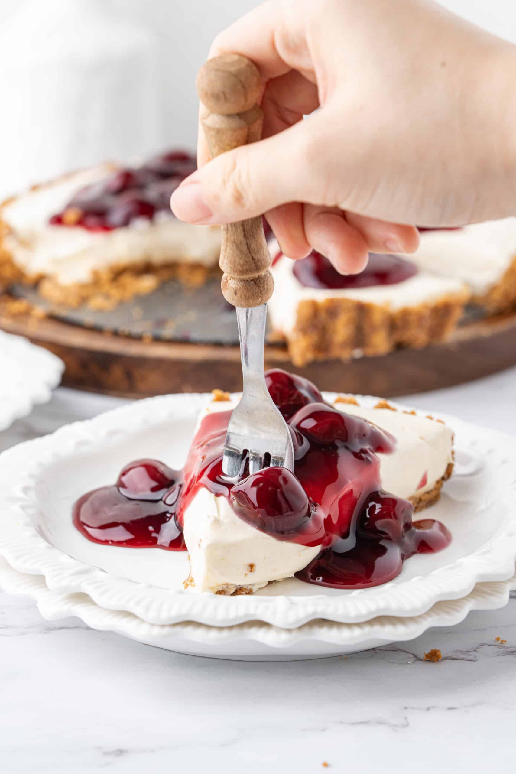 Hand holding a fork over a slice of no-bake cheesecake topped with cherry pie filling on a white dessert plate.