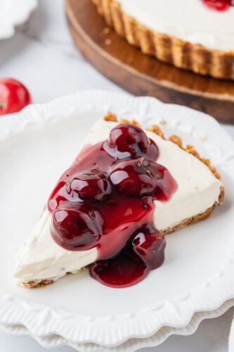 Slice of no-bake cheesecake with graham cracker crust topped with cherry pie filling on a white dessert plate.