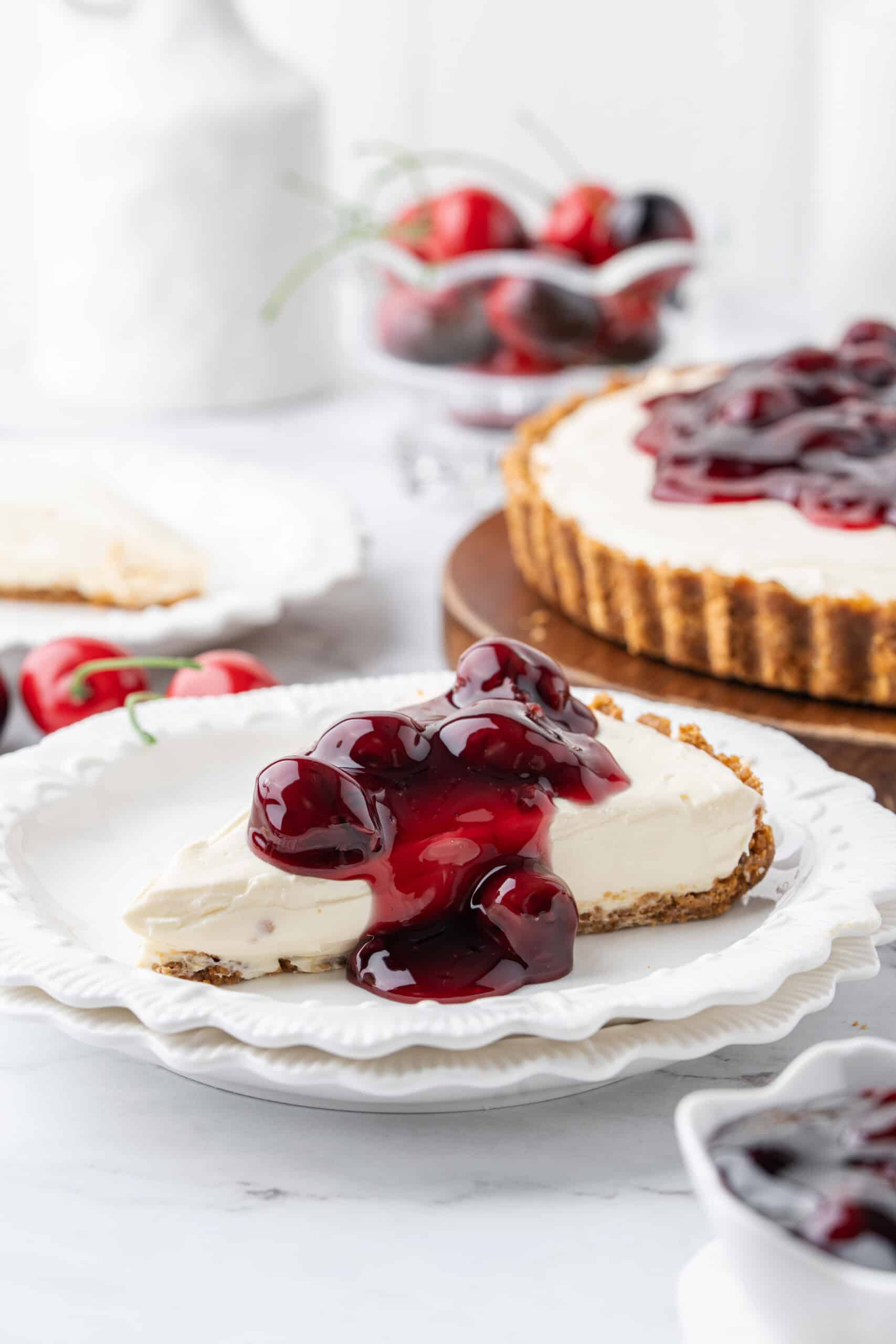 cherry no bake cheesecake recipe Slice of no-bake cheesecake with graham cracker crust and cherry pie filling served on a white dessert plate, with the cheesecake in the background.