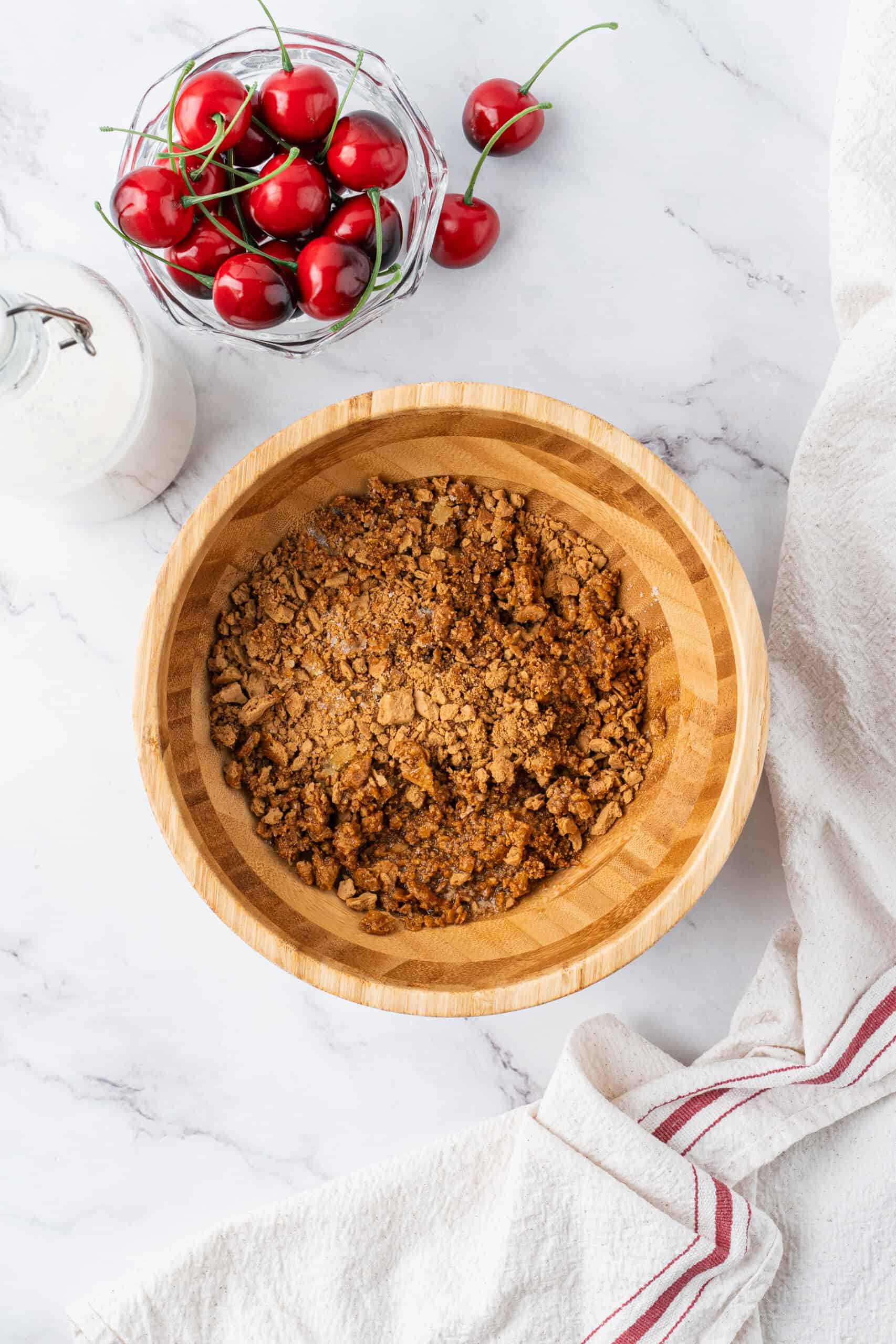 Mixing graham cracker crumbs, melted butter, and sugar in a wooden bowl with fresh cherries and a bottle of milk nearby.