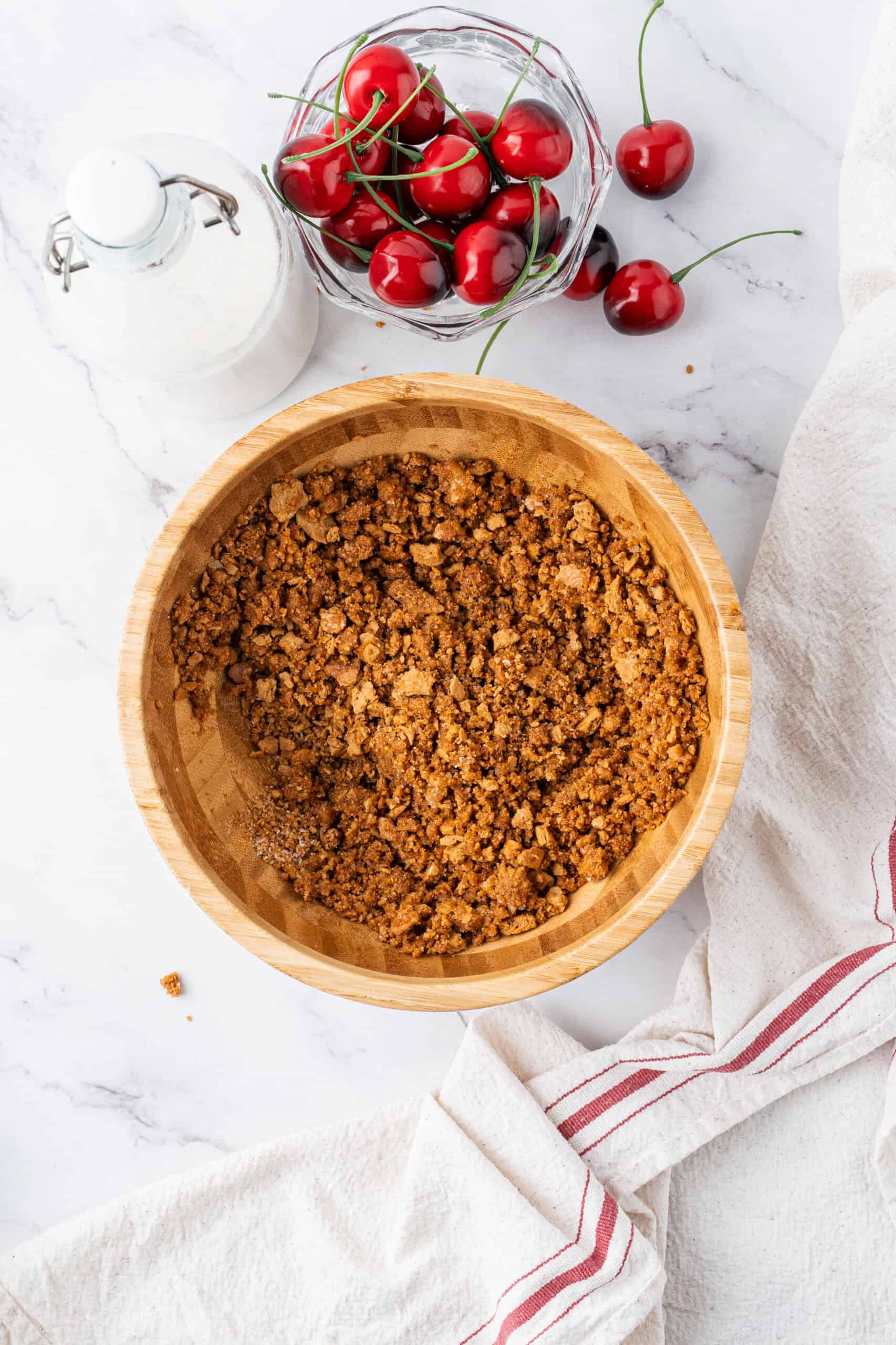 Graham cracker crust mixture combined in a wooden bowl with fresh cherries and a bottle of milk on the counter.