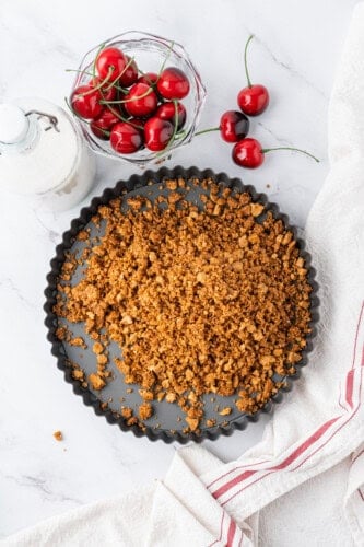 Graham cracker crust mixture spread into a pie pan, with fresh cherries and a bottle of milk nearby.