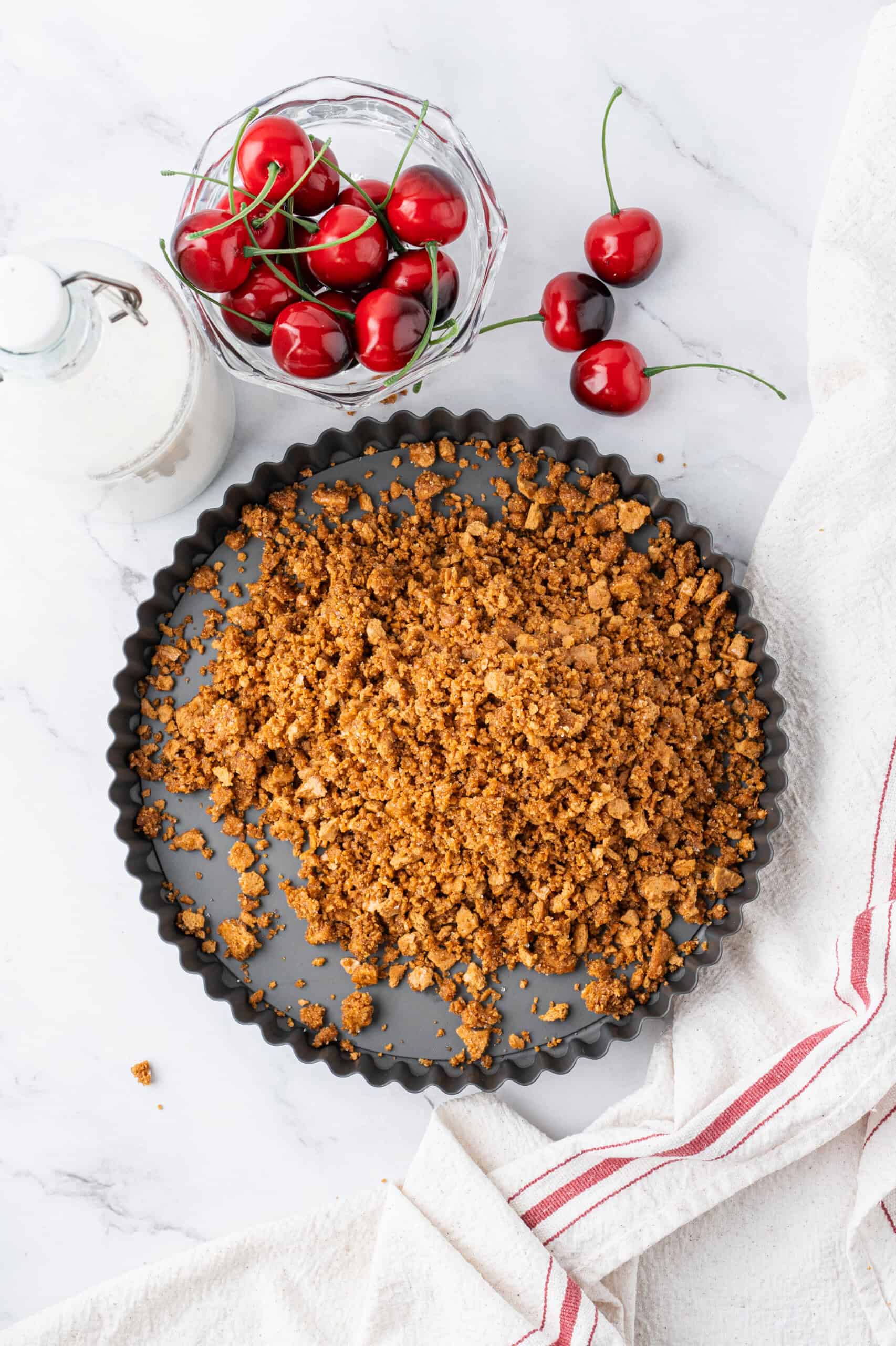 Graham cracker crust mixture spread into a pie pan, with fresh cherries and a bottle of milk nearby.