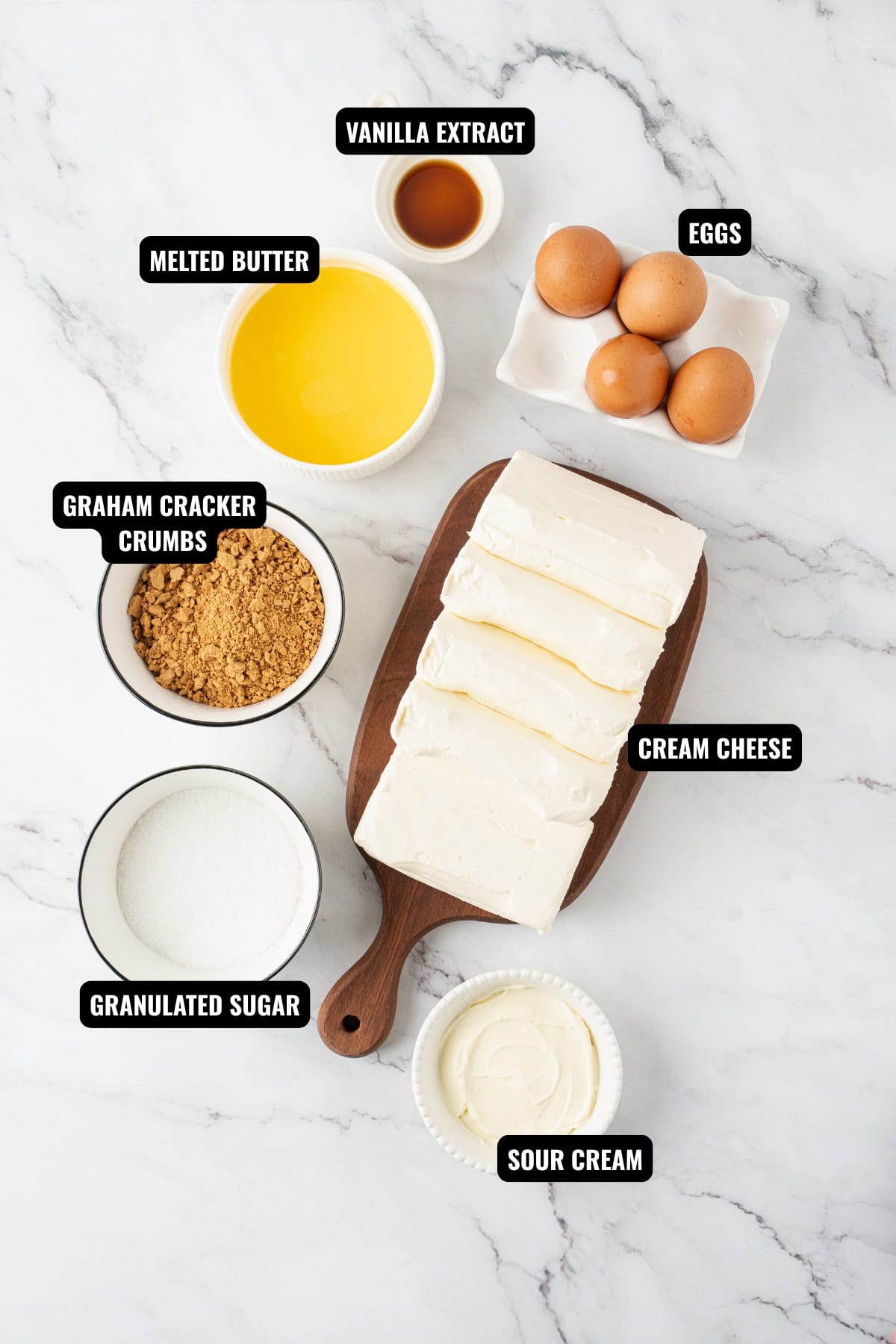 ingredients for cheesecake recipe including graham cracker crumbs, cream cheese, melted butter, eggs, sugar, sour cream, and vanilla extract.