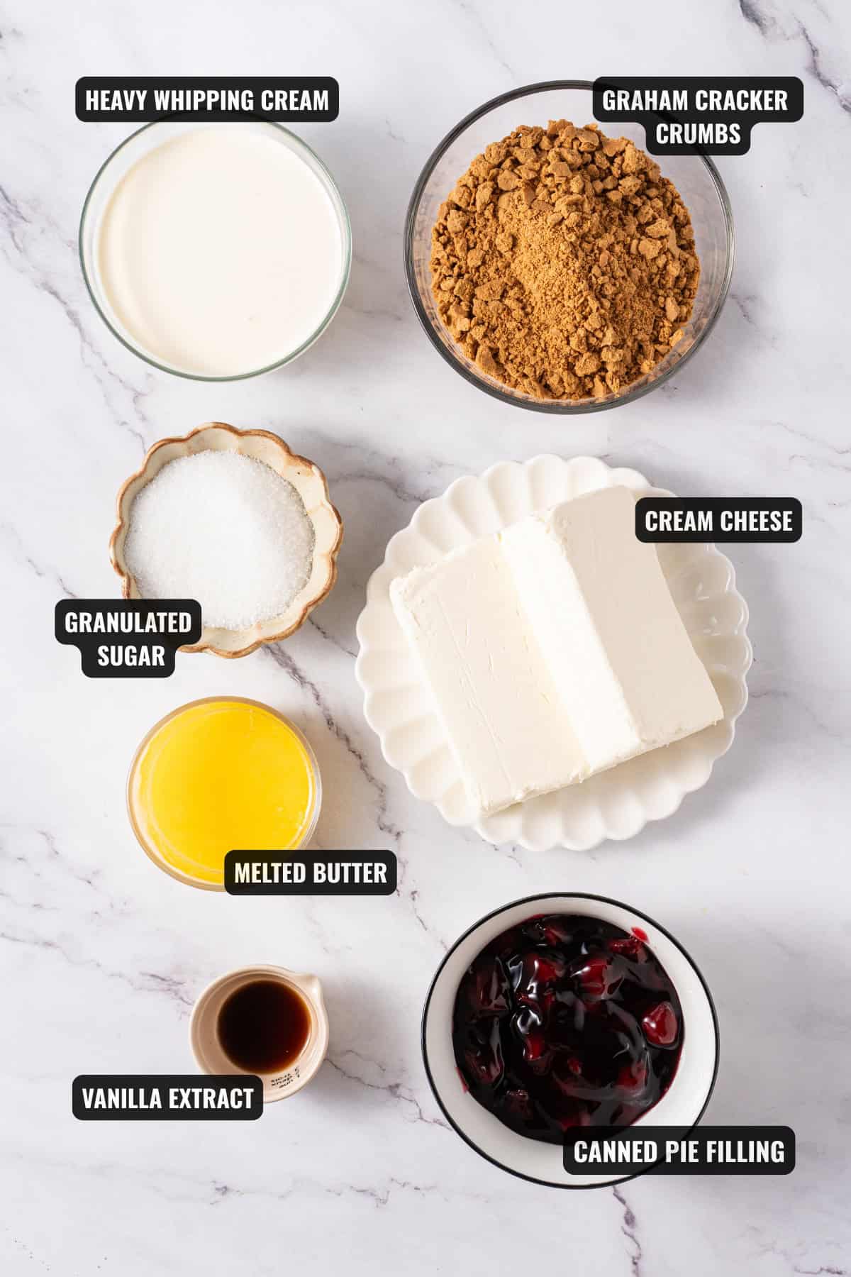 ingredients for no-bake cheesecake recipe including graham cracker crumbs, heavy whipping cream, cream cheese, granulated sugar, melted butter, canned pie filling, and vanilla extract.