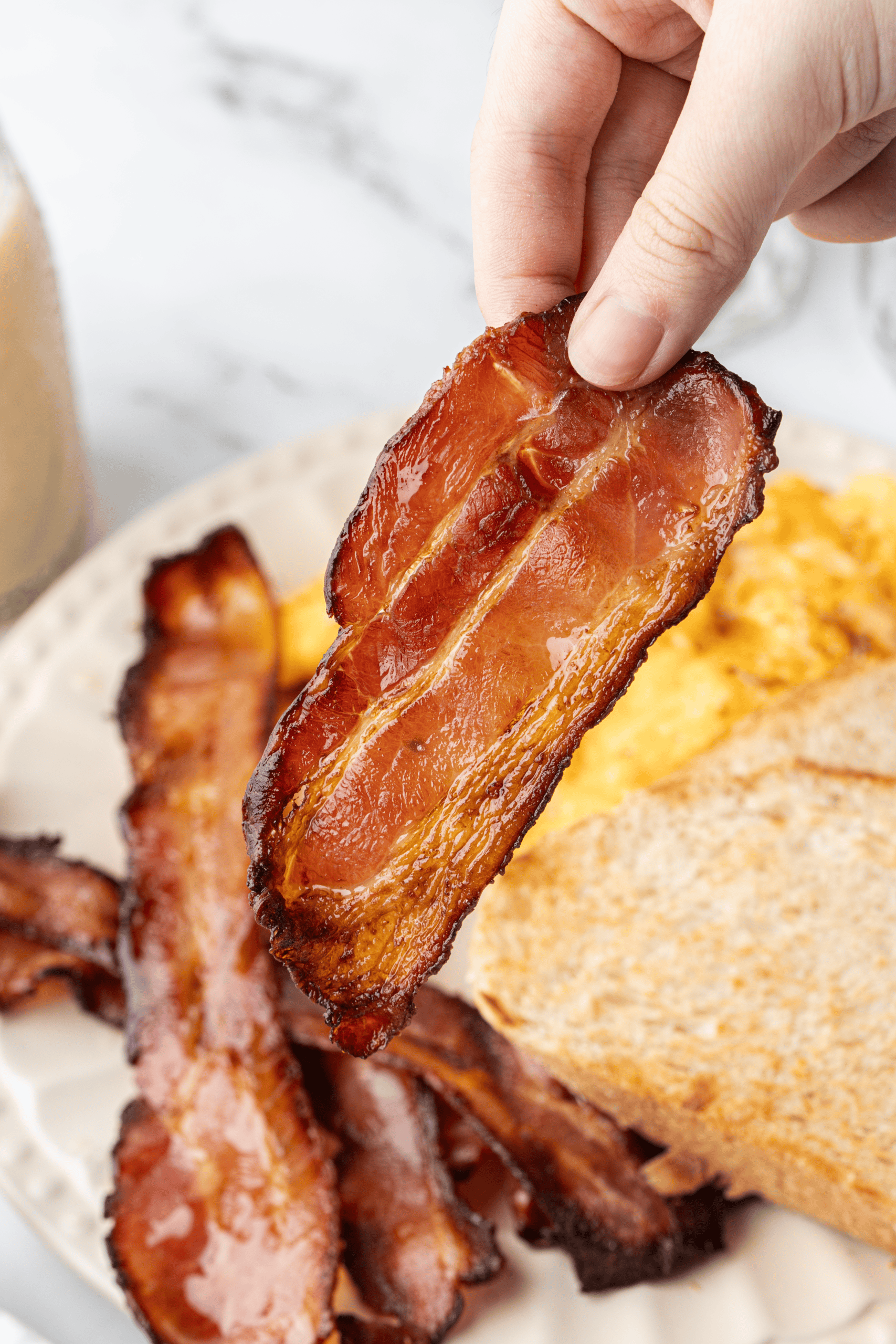 Crispy oven-baked bacon held over a breakfast plate.