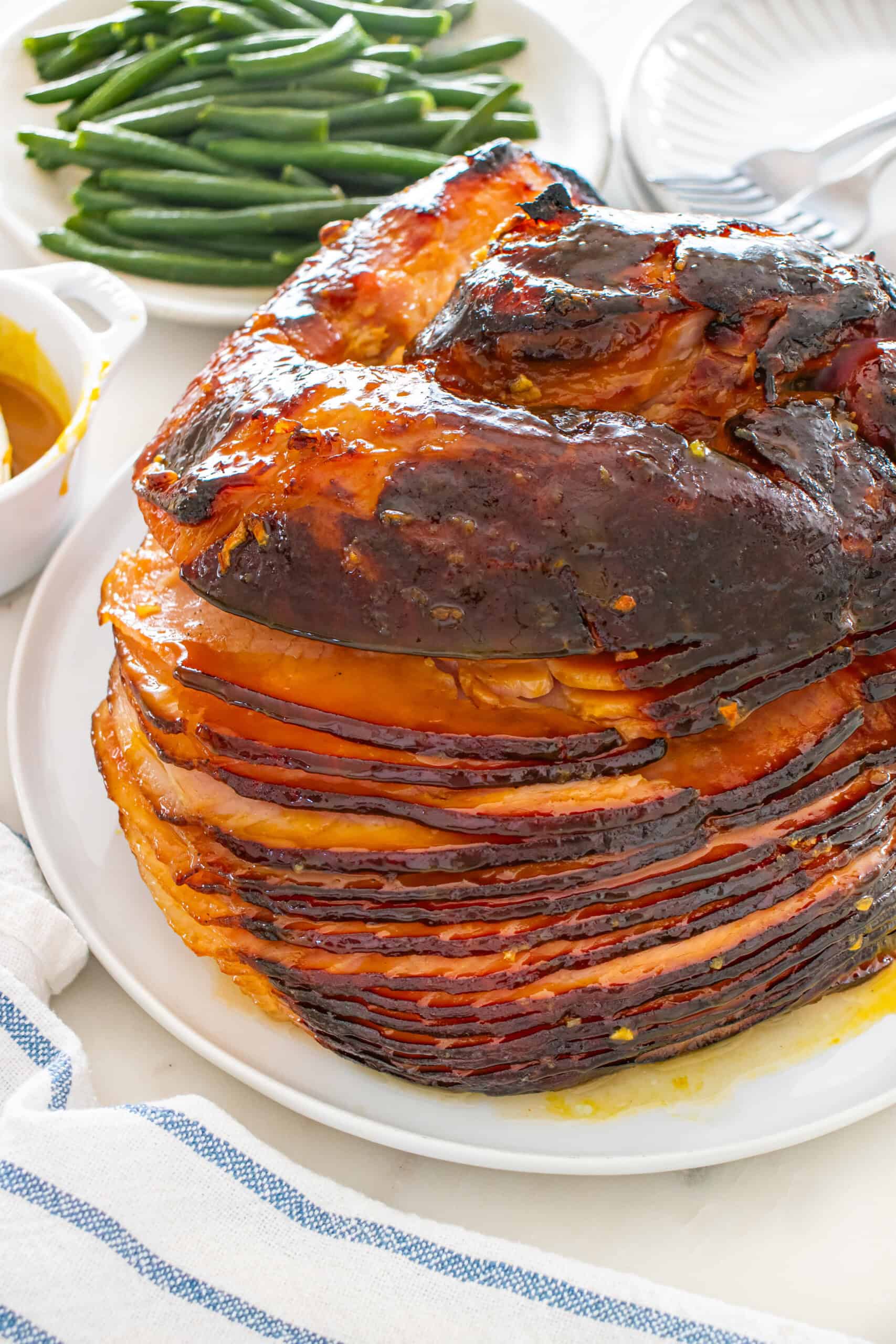Close-up of glazed baked ham on a serving platter with green beans and extra glaze.