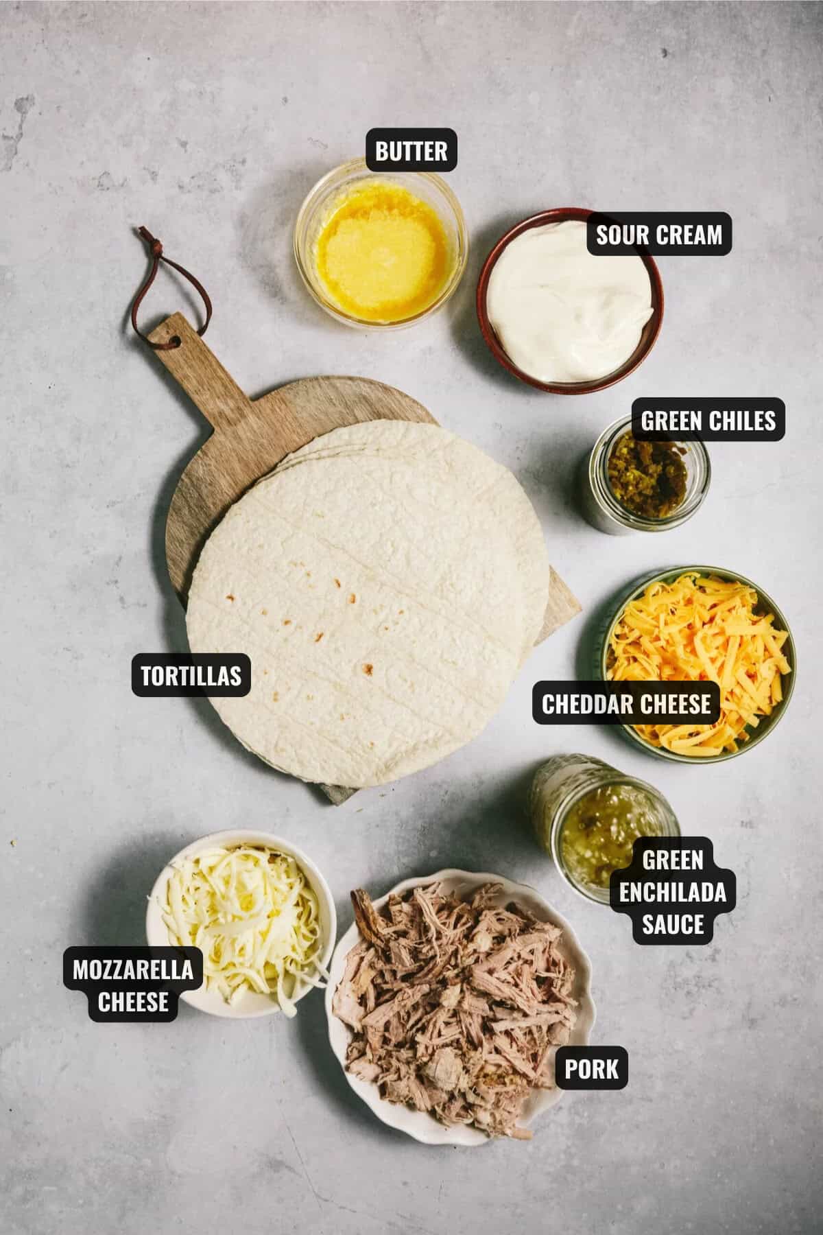 ingredients for pork enchiladas including pork, tortillas, green enchilada sauce, green chiles, sour cream, butter, cheddar and mozzarella cheese.