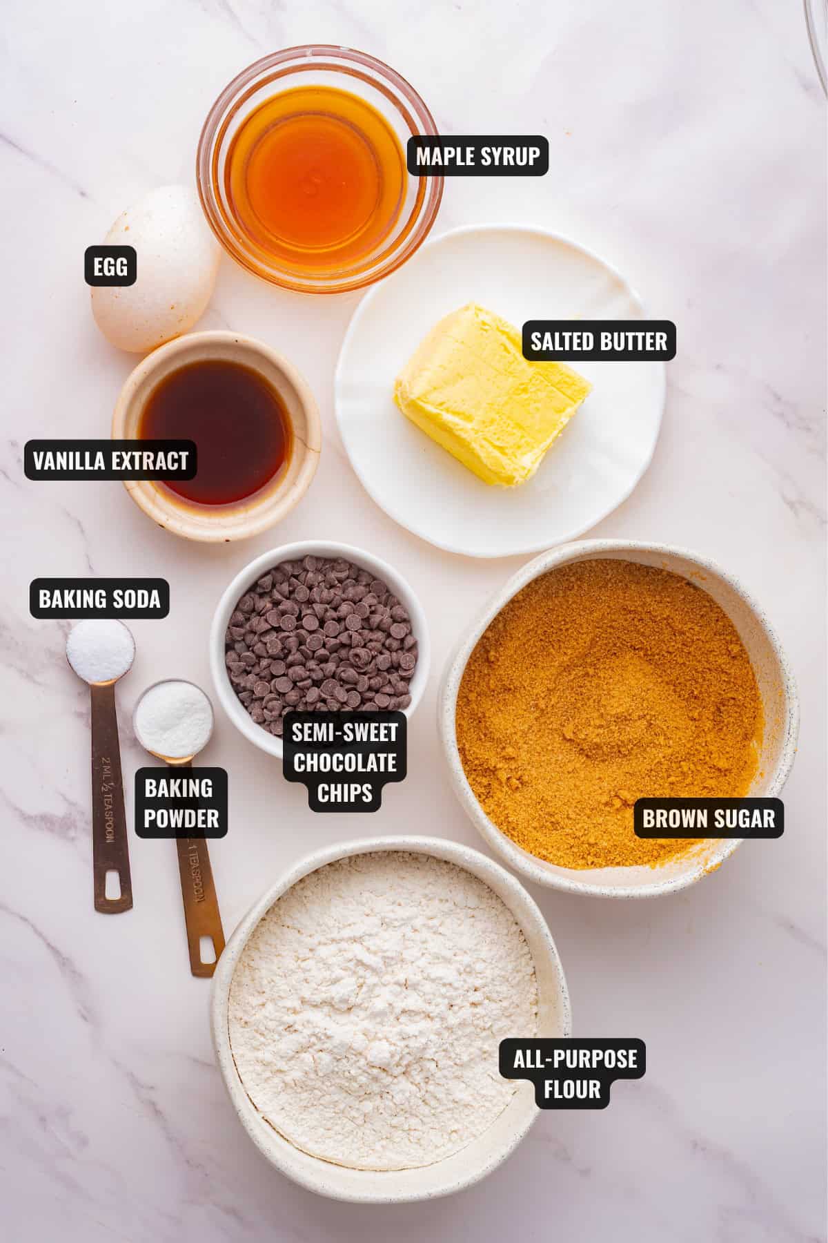 ingredients for maple cookies recipe including maple syrup, egg, butter, flour, sugar, baking soda, baking powder, chocolate chips, and vanilla extract.