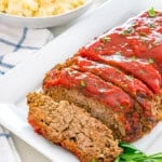 Sliced meatloaf with glaze and parsley, served with mashed potatoes and green beans.