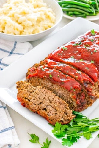 Sliced meatloaf with glaze and parsley, served with mashed potatoes and green beans.