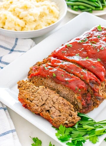 Sliced meatloaf with glaze and parsley, served with mashed potatoes and green beans.