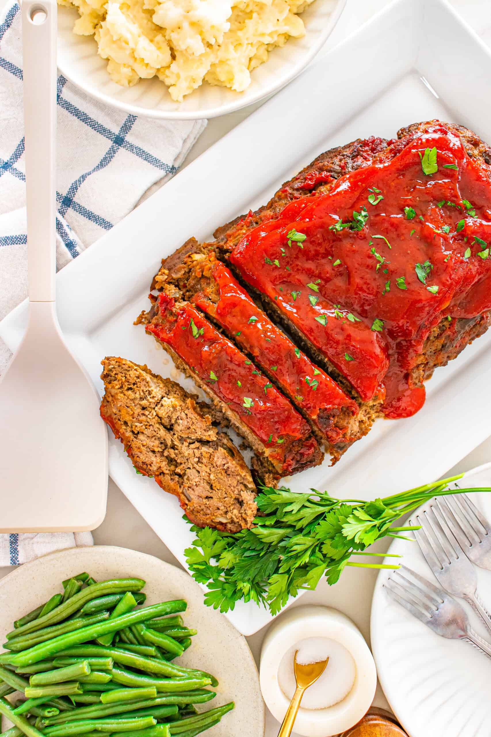 Sliced moist meatloaf with glaze and parsley on a serving platter with mashed potatoes and green beans.