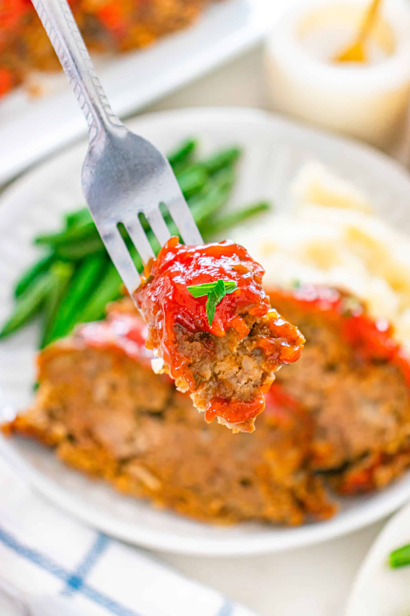 Bite of glazed moist meatloaf on a fork with green beans and mashed potatoes in the background.