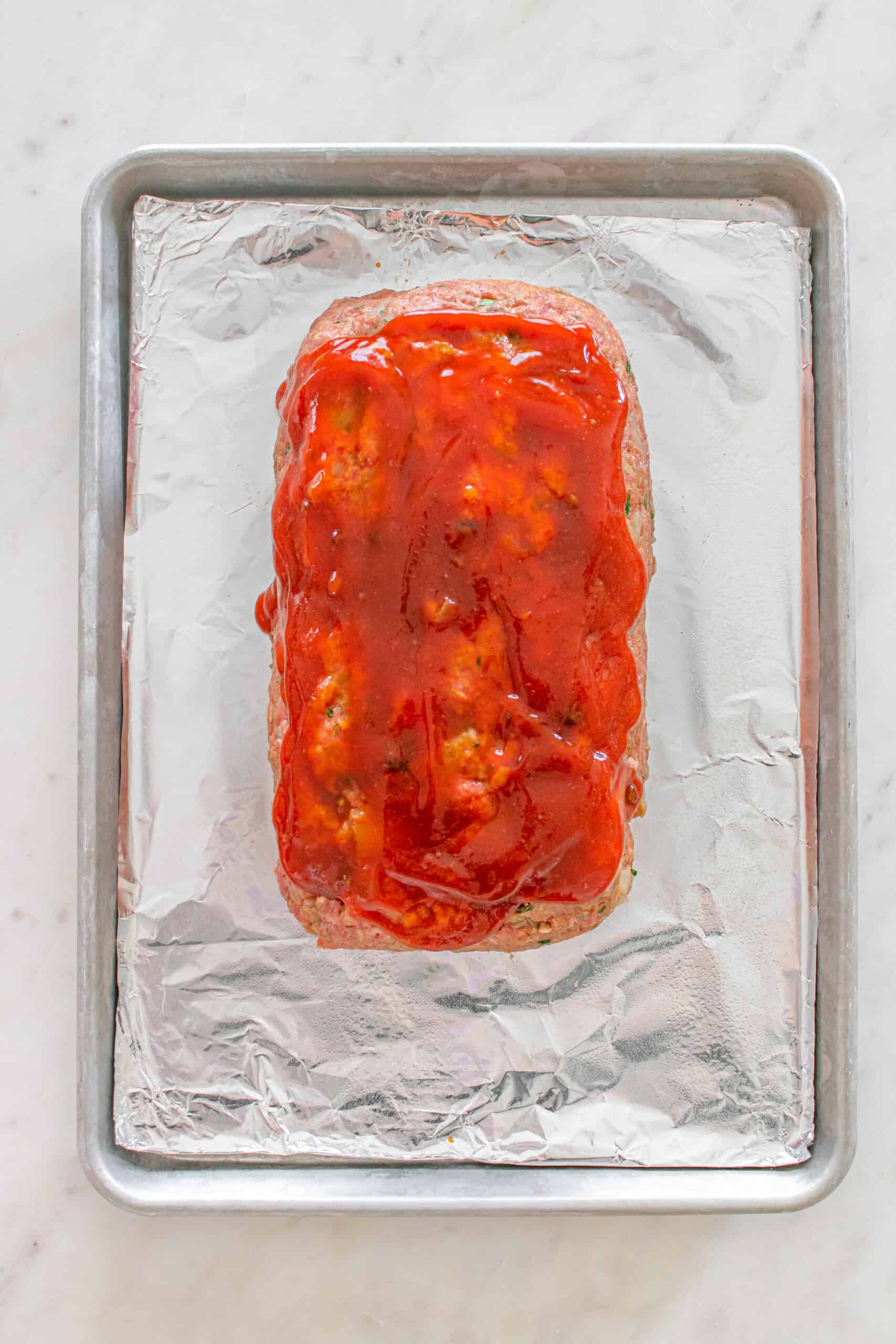 Uncooked meatloaf topped with glaze on a foil-lined baking sheet.