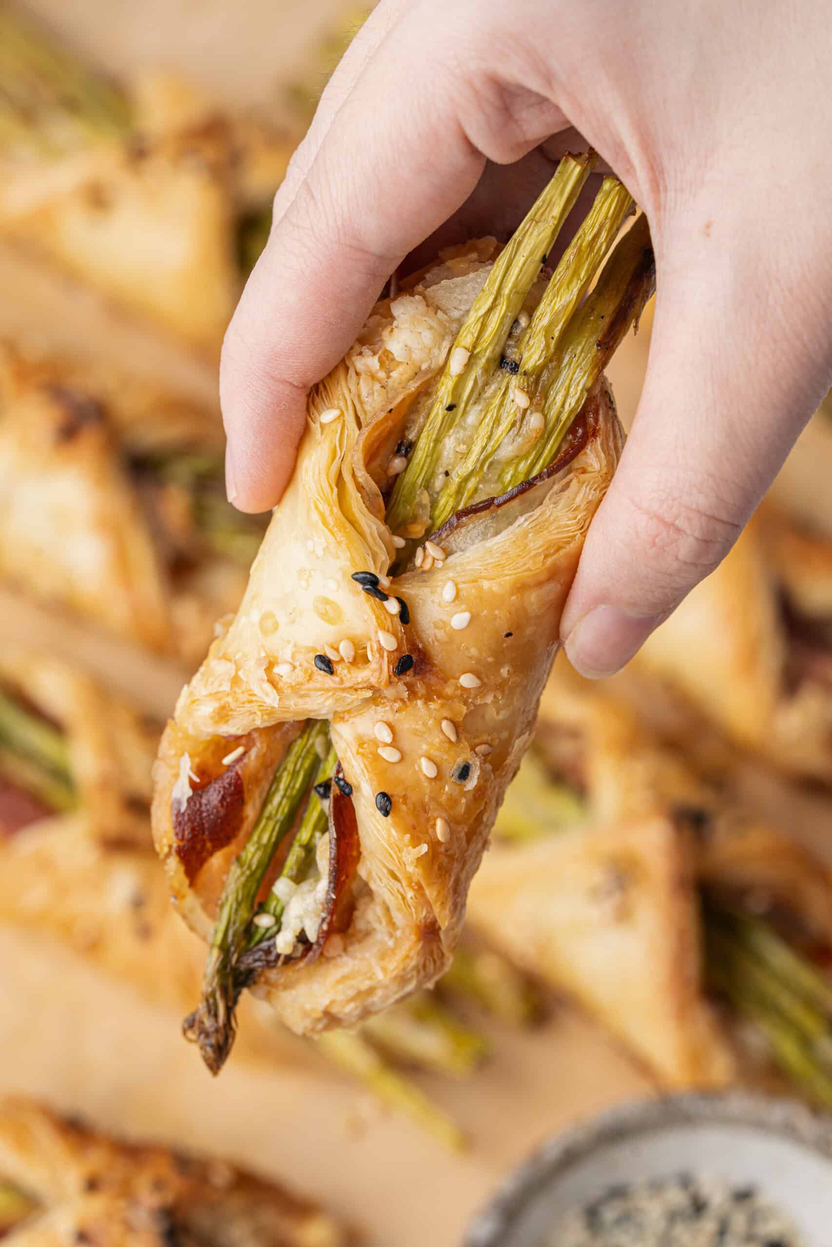 Hand holding a golden asparagus bundle with pancetta and sesame seeds.