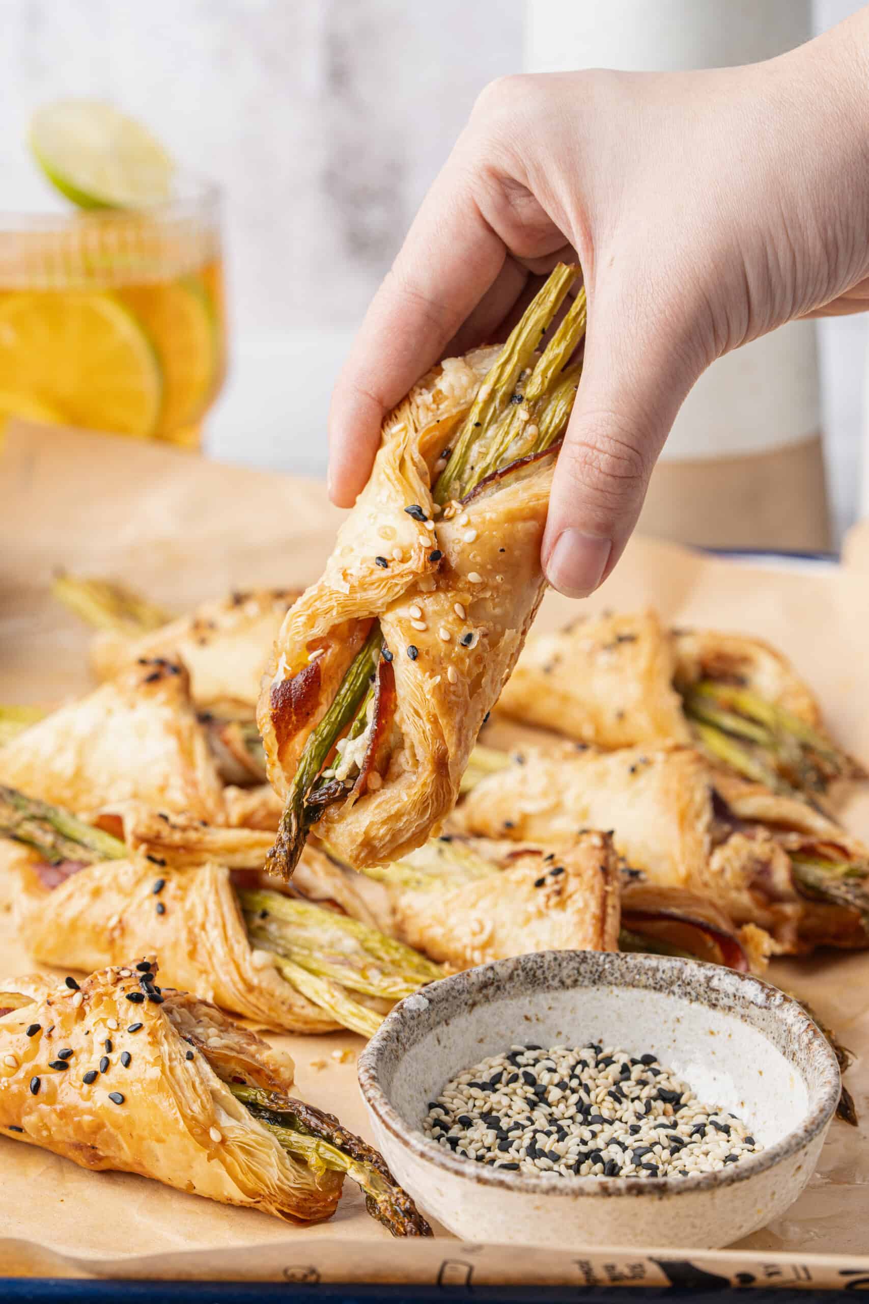 Hand holding a baked asparagus puff pastry bundle with pancetta and sesame seeds.
