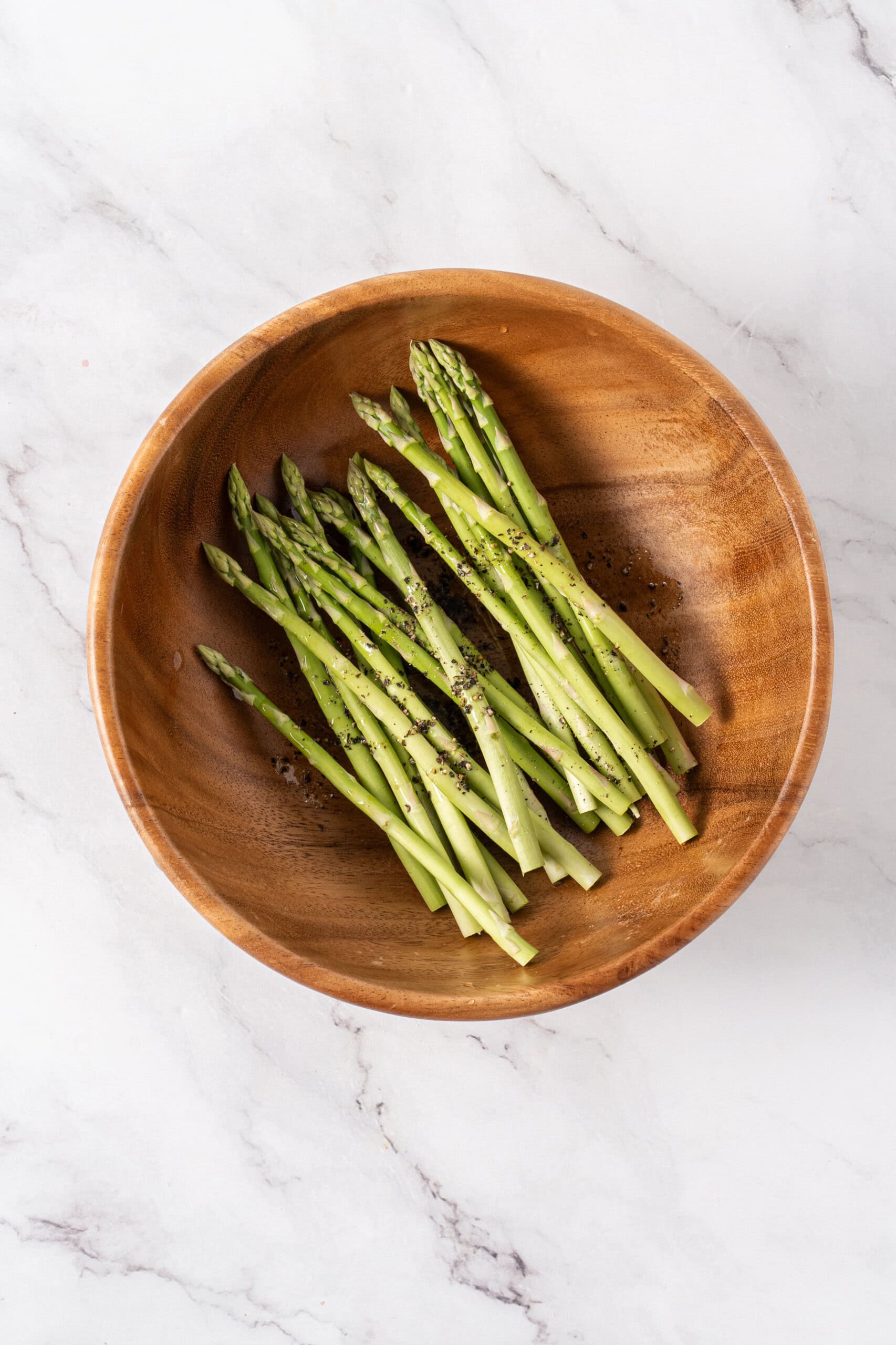 Asparagus spears seasoned with olive oil and freshly ground black pepper in a wooden bowl.