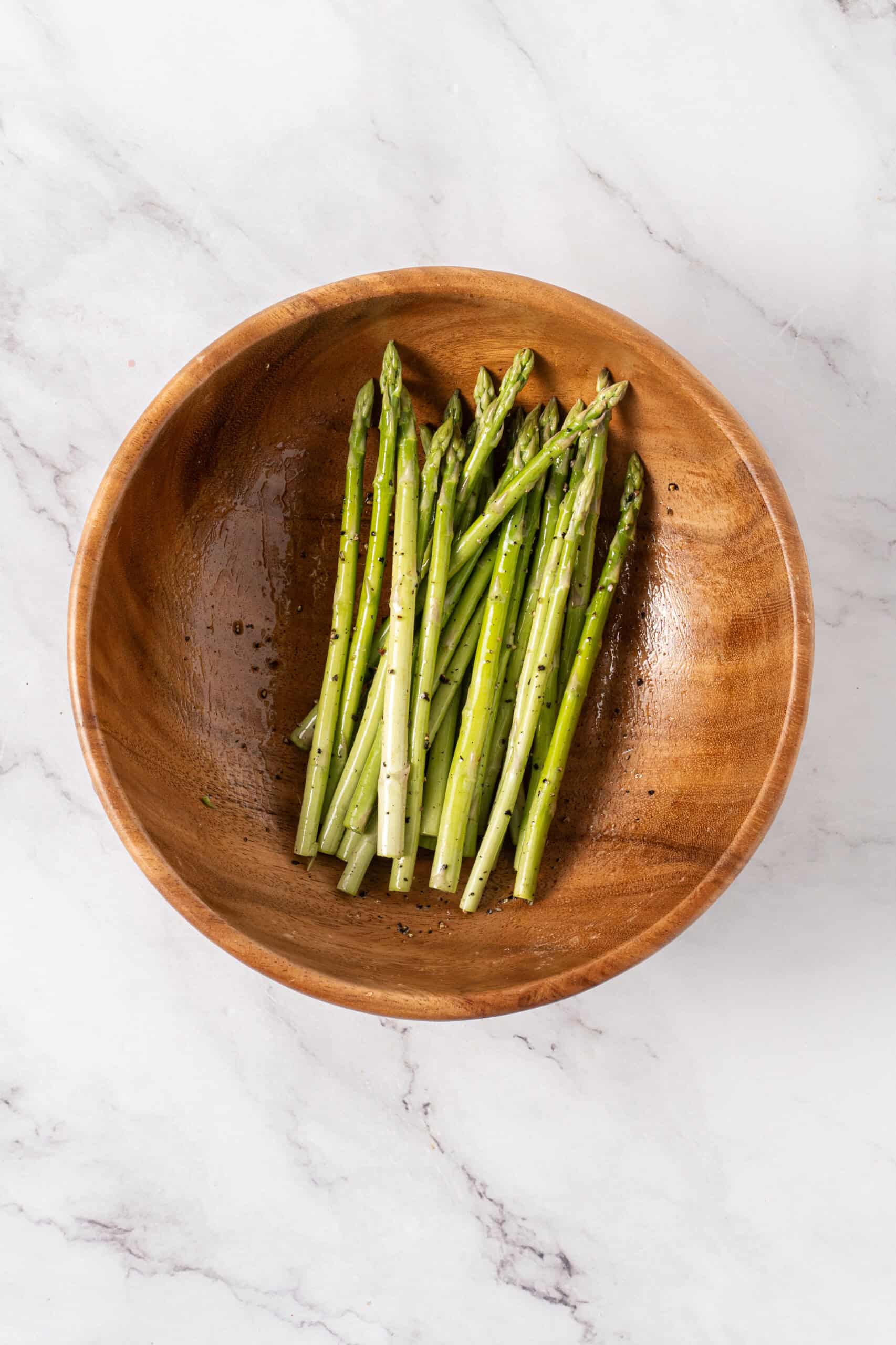 Asparagus spears coated with olive oil and black pepper in a wooden bowl.