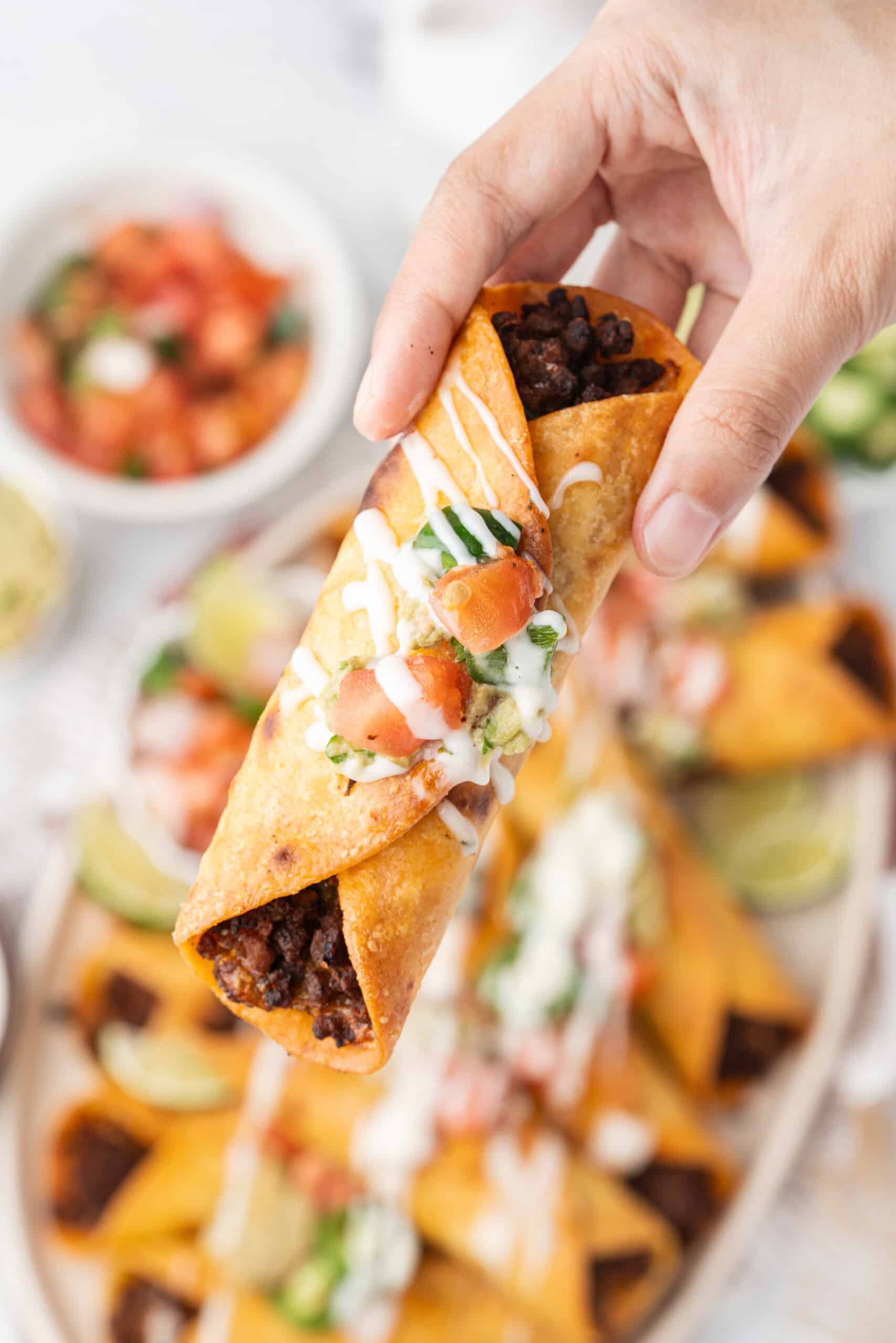 A hand holding a crispy taquito filled with seasoned ground beef and topped with guacamole, pico de gallo, sliced jalapeño, and crema, with a platter of taquitos blurred in the background.
