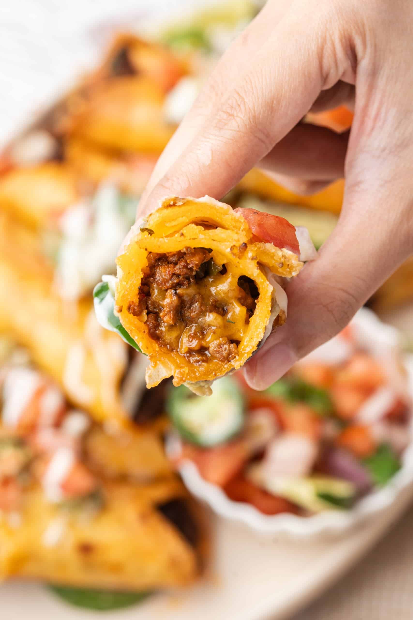 Close-up of a crispy taquito filled with seasoned ground beef and melted cheese, held above a platter of taquitos and fresh toppings.