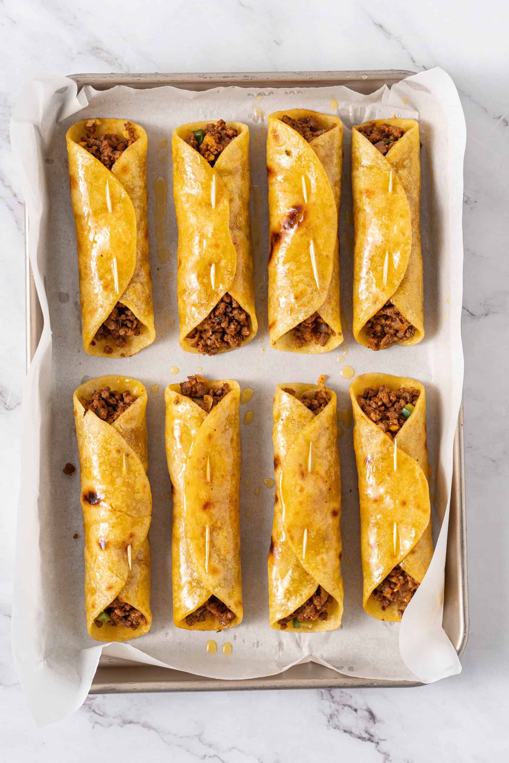 Taquitos brushed with butter on a parchment-lined baking sheet.