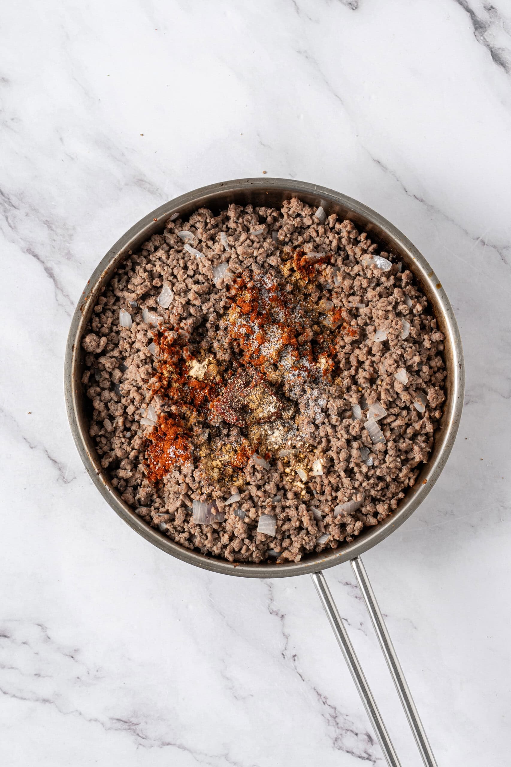 Ground beef and onions topped with seasonings in a skillet.