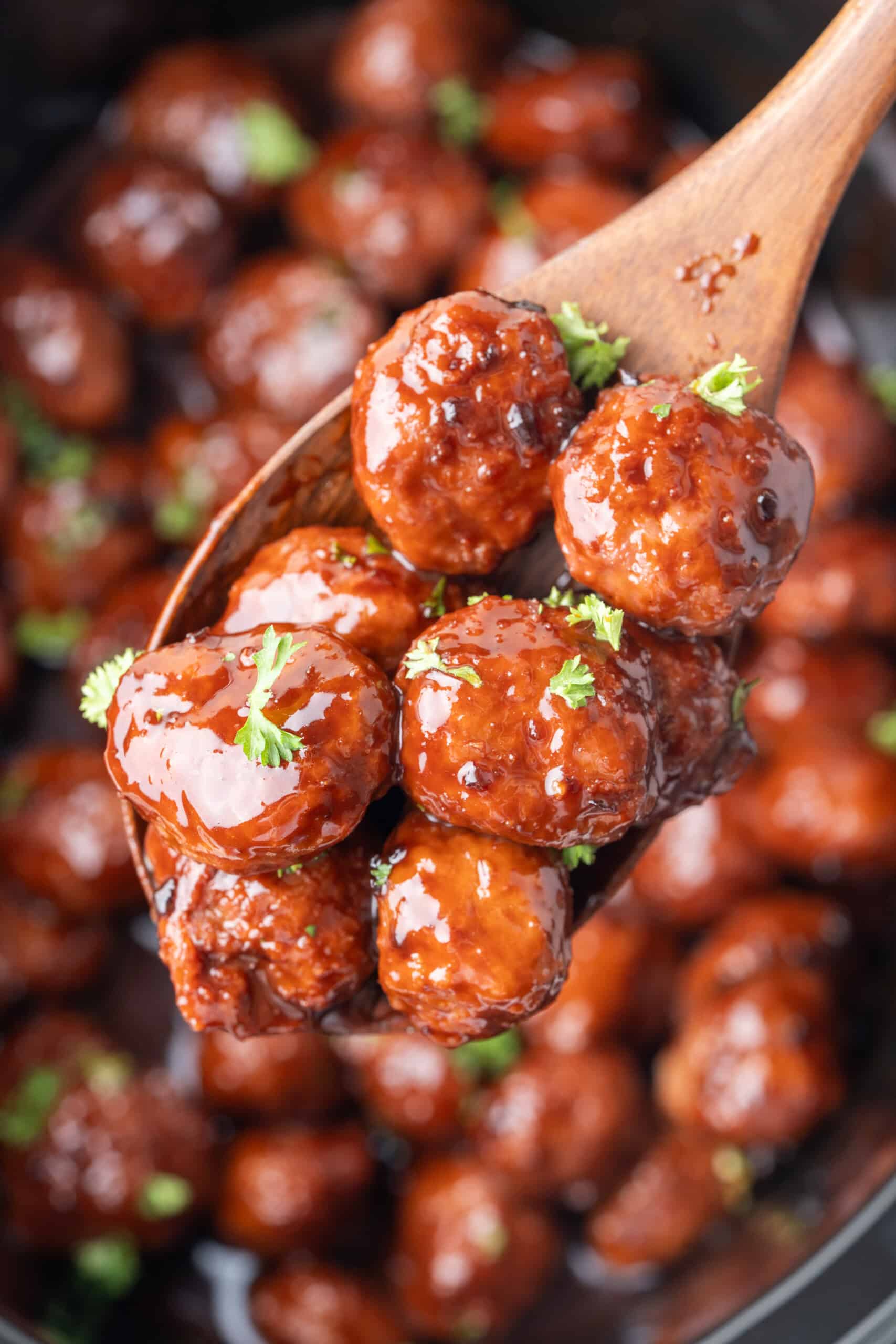 Grape jelly meatballs coated in glossy sauce and garnished with fresh parsley on a wooden spoon.