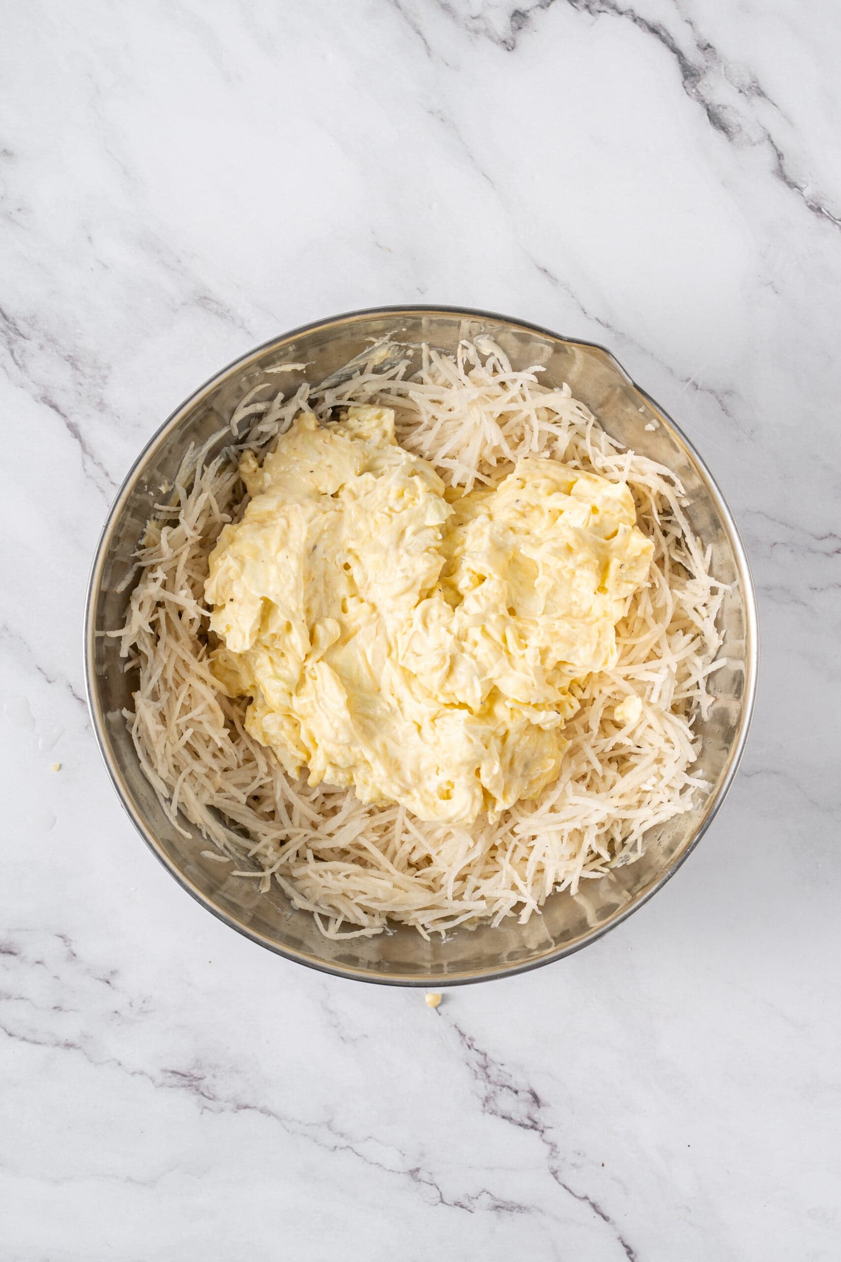 Shredded hash browns topped with the creamy cheese mixture in a large mixing bowl.