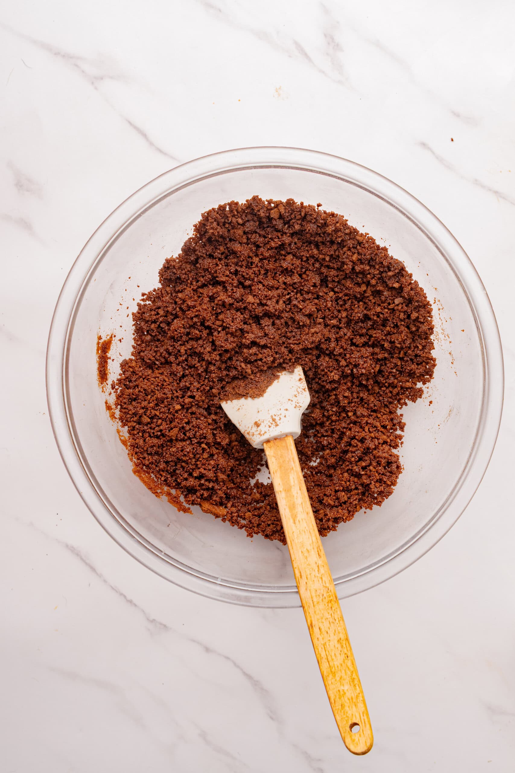 Chocolate cookie crumbs mixed with melted butter in a glass bowl with a spatula.