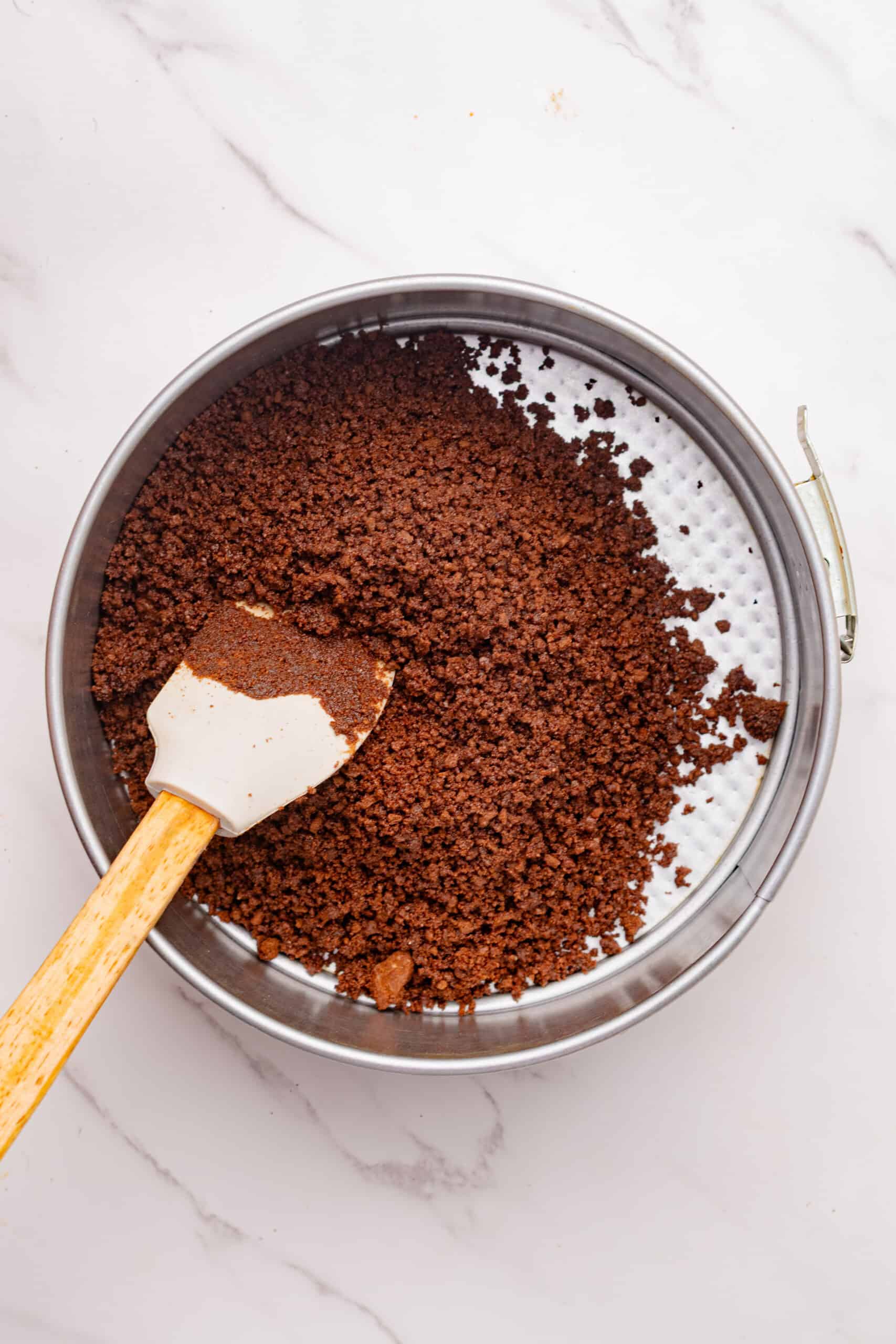 Chocolate cookie crumb mixture spread into a springform pan with a spatula.