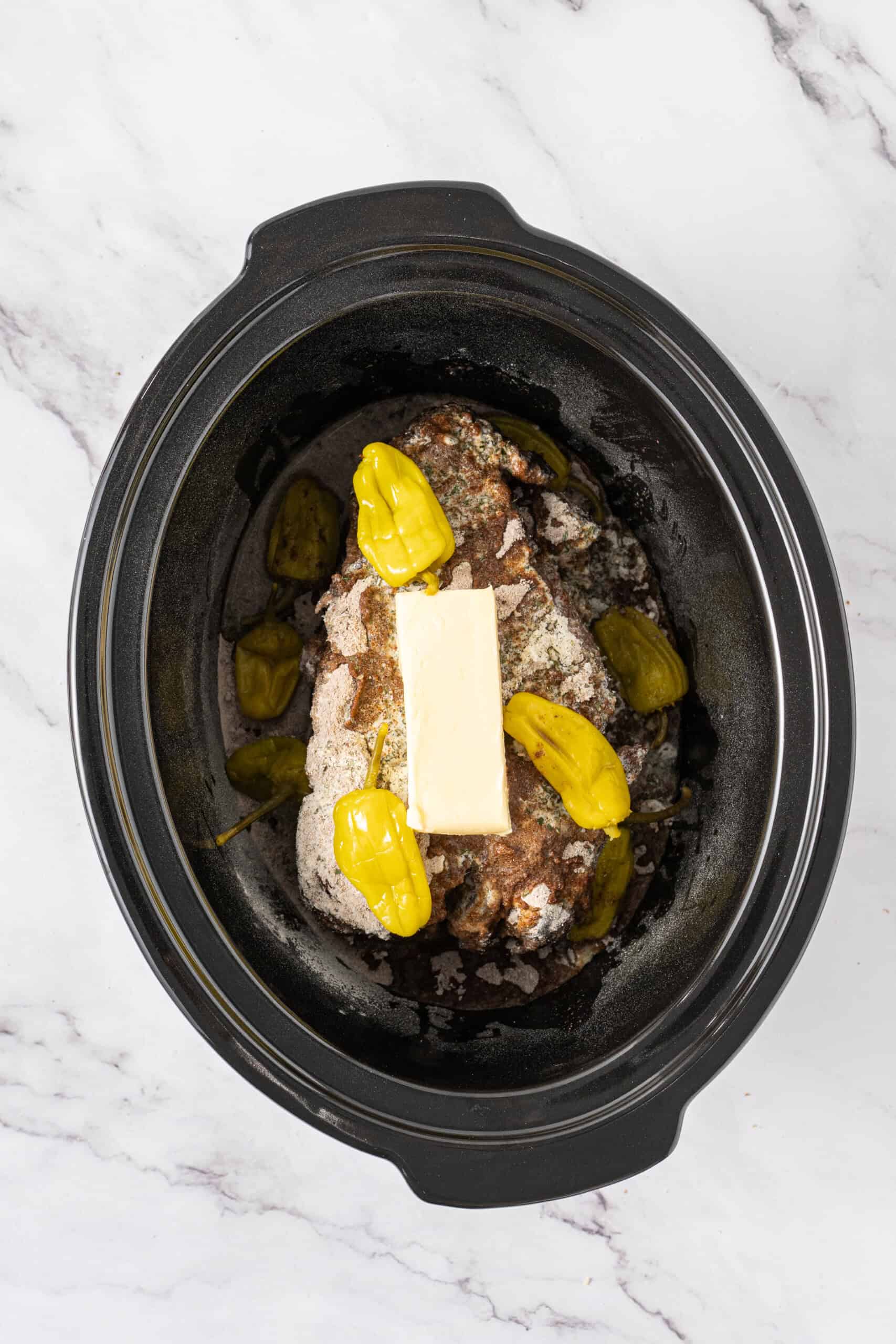 Seared beef roast topped with seasoning, butter, and pepperoncini in a slow cooker.