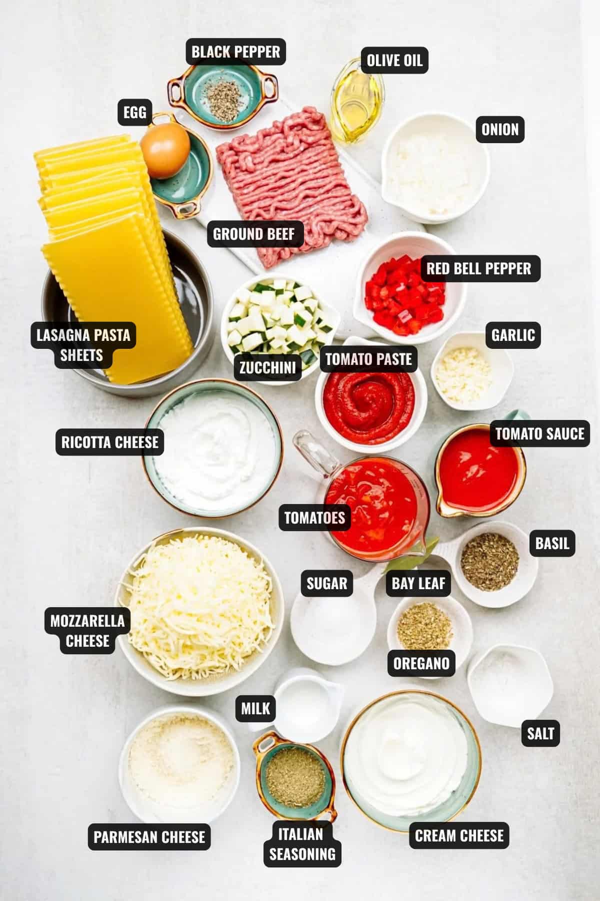 ingredients for lasagna cups recipe including lasagna pasta sheets, egg, black pepper, olive oil, onion, ground beef, zucchini, tomato paste, red bell pepper, garlic, tomato paste, tomato sauce, tomatoes, ricotta cheese, mozzarella cheese, parmesan cheese, cream cheese, italian seasoning, milk, sugar, bay leaf, dried basil, dried oregano, and salt.