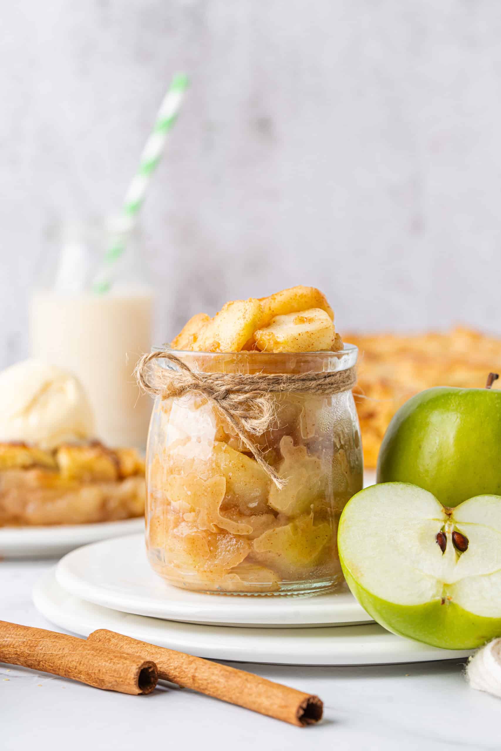 Apple Pie Filling with cinnamon-coated apples in a glass jar beside fresh green apples and cinnamon sticks.