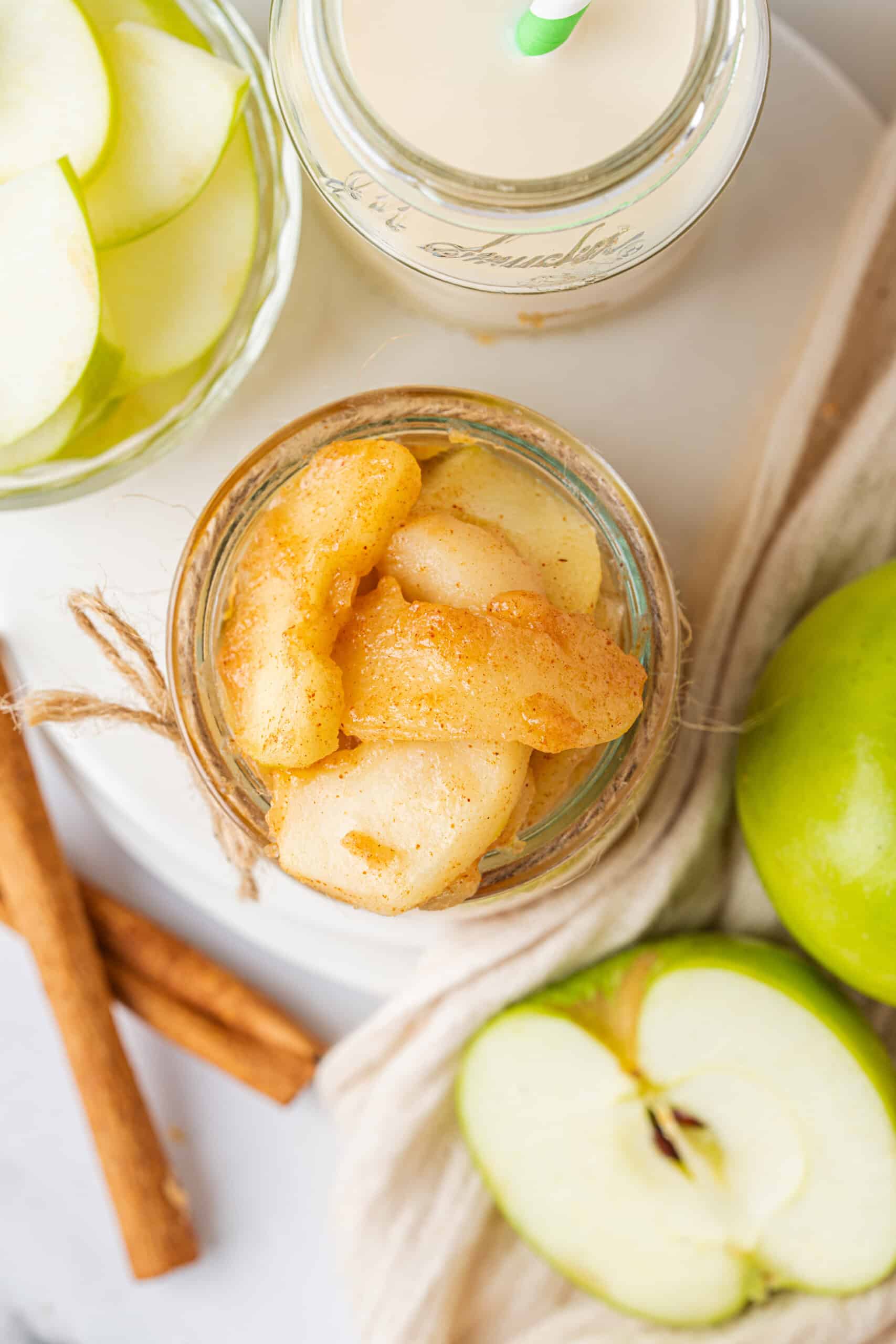 Apple Pie Filling with cinnamon-coated apple slices in a glass jar surrounded by fresh green apples and cinnamon sticks.