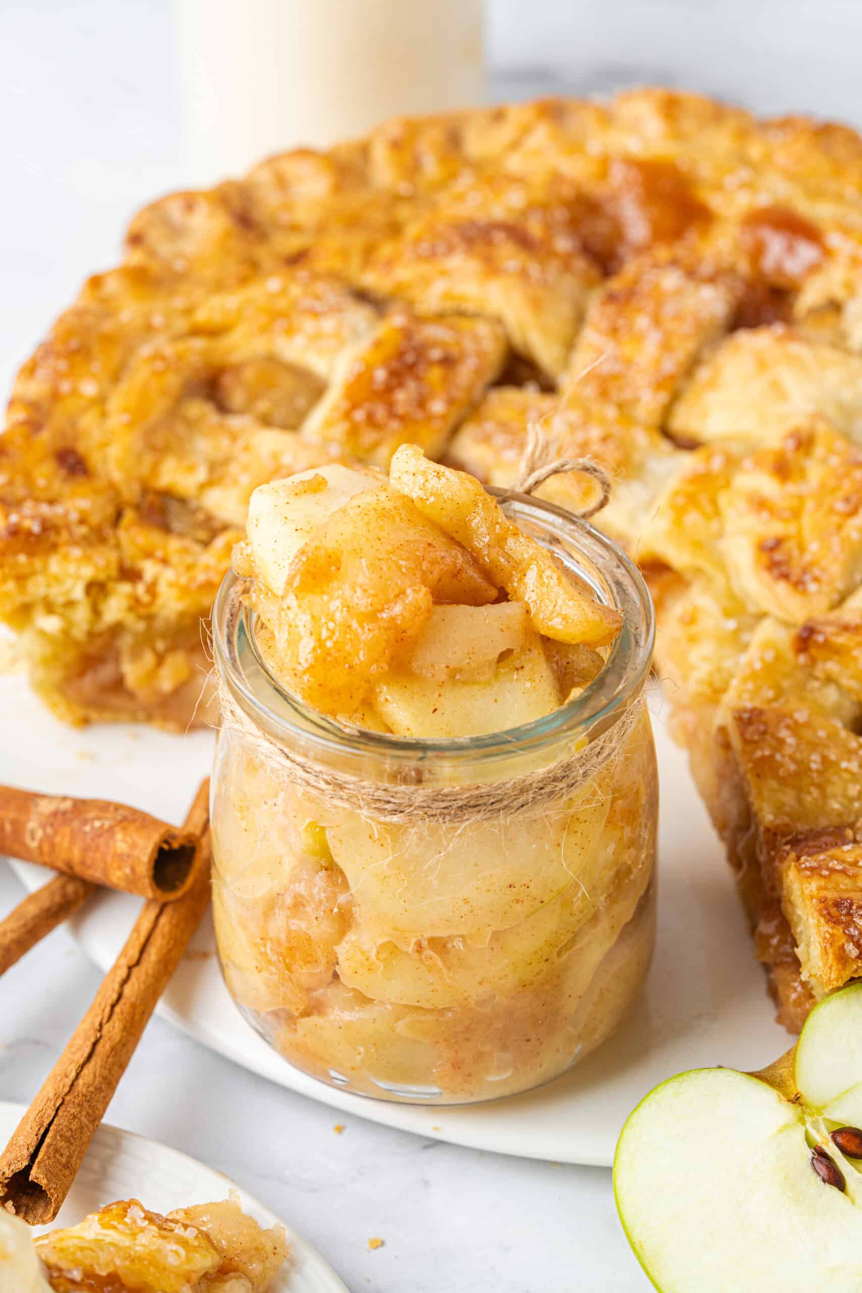 Homemade Apple Pie Filling with tender cinnamon-coated apple slices in a glass jar, surrounded by golden apple pie and fresh apples.
