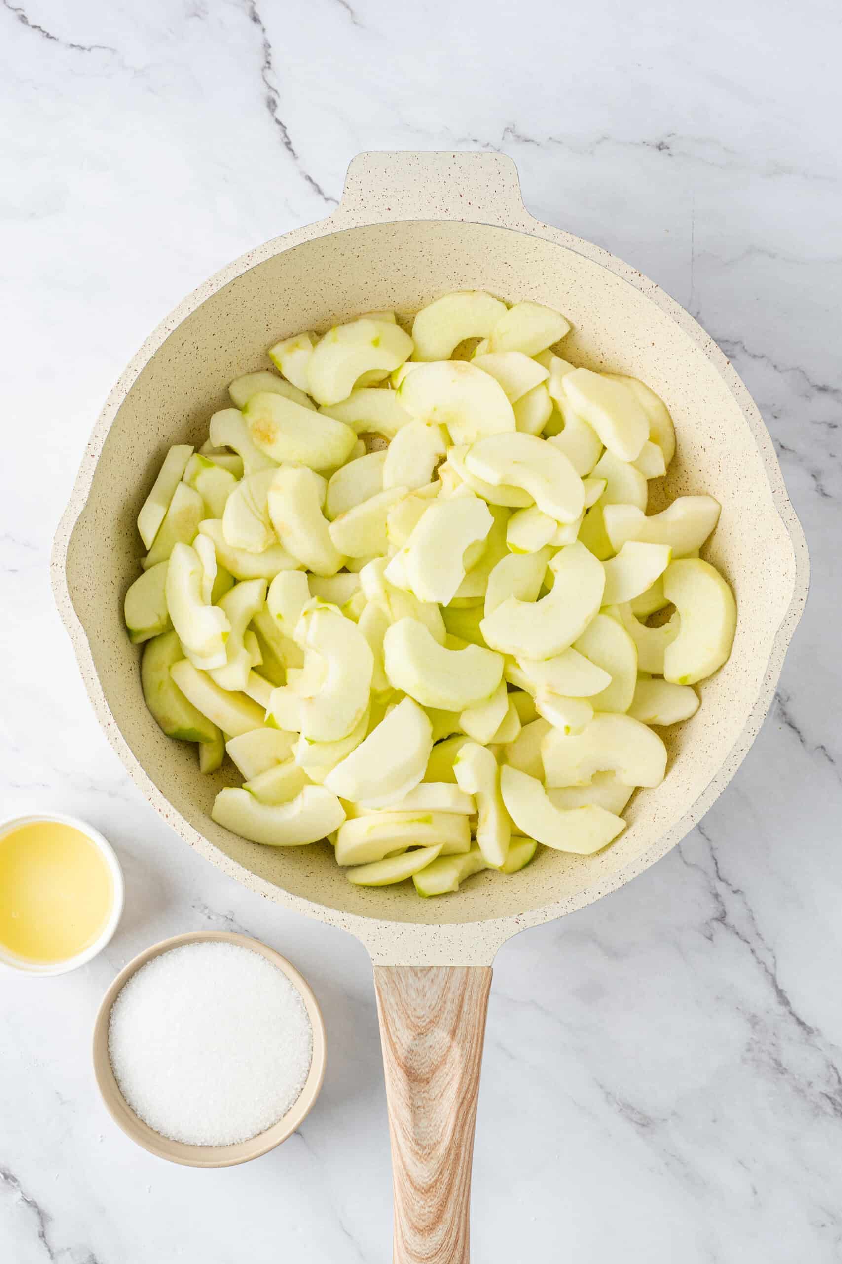 Peeled and sliced apples in a cream-colored skillet, with bowls of sugar and lemon juice alongside.