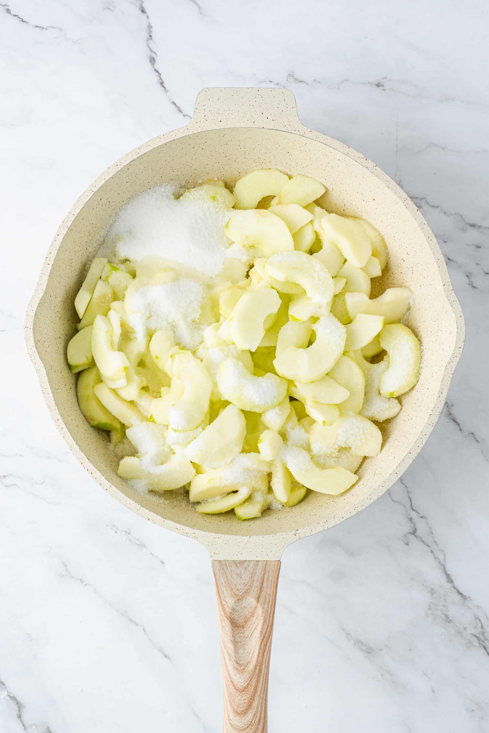 Peeled and sliced apples sprinkled with granulated sugar in a cream-colored skillet.