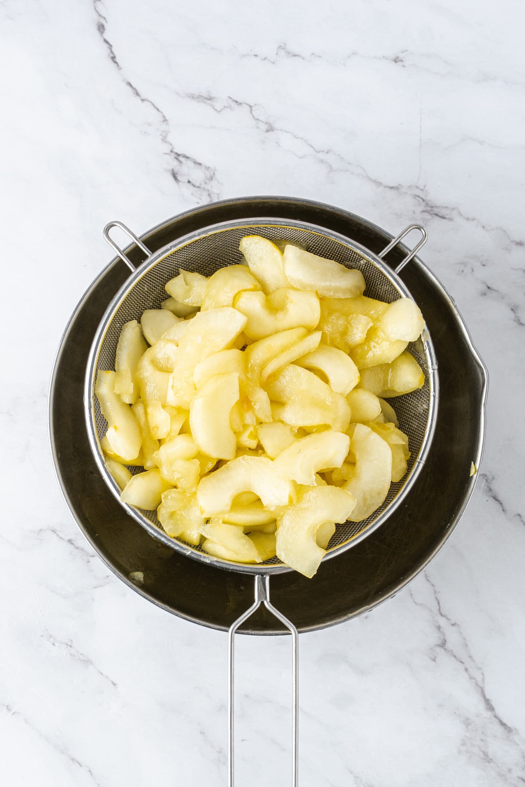 Cooked apple slices draining in a fine-mesh strainer over a metal bowl.