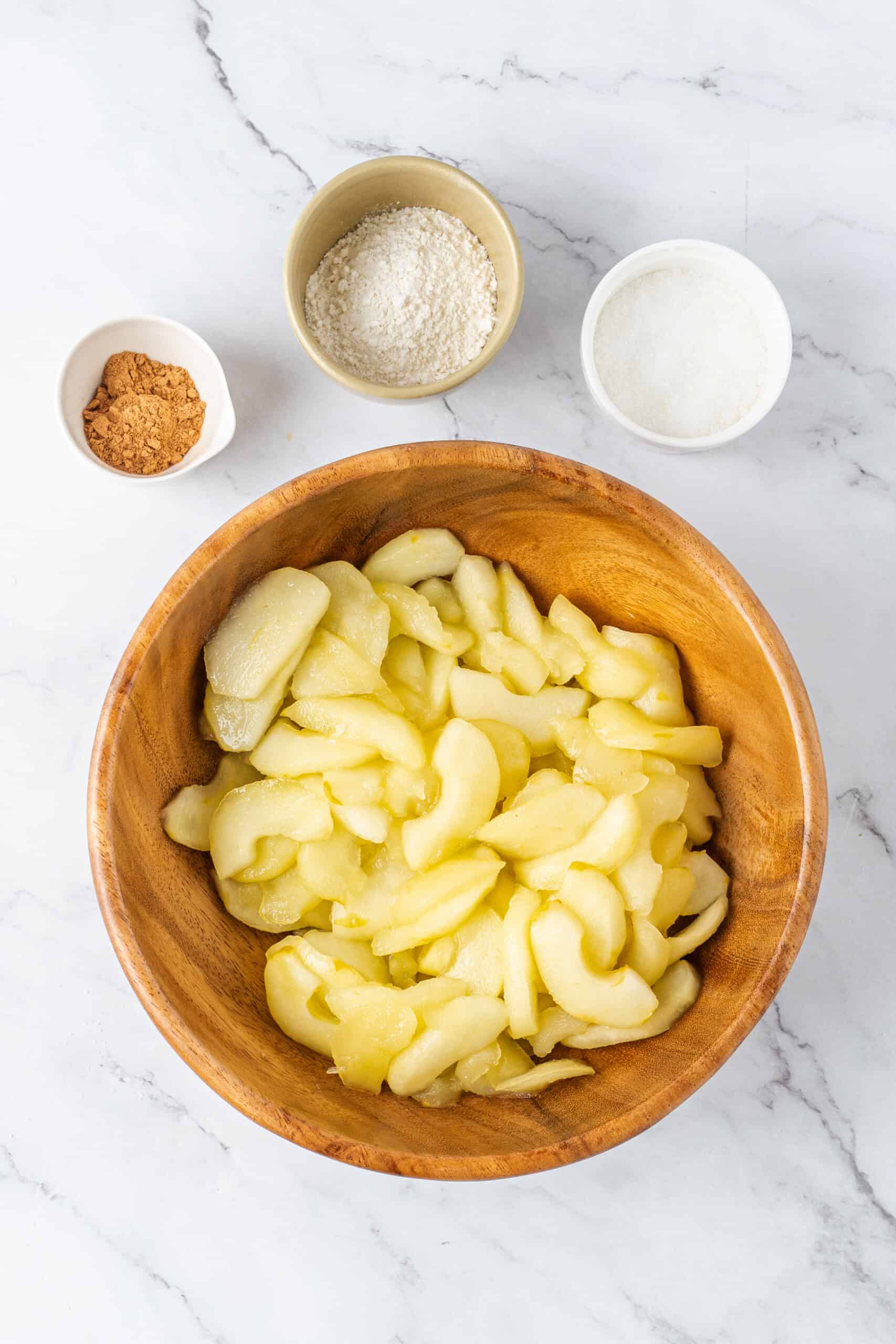 Cooked apple slices in a wooden bowl with cinnamon, flour, and sugar in small bowls above.