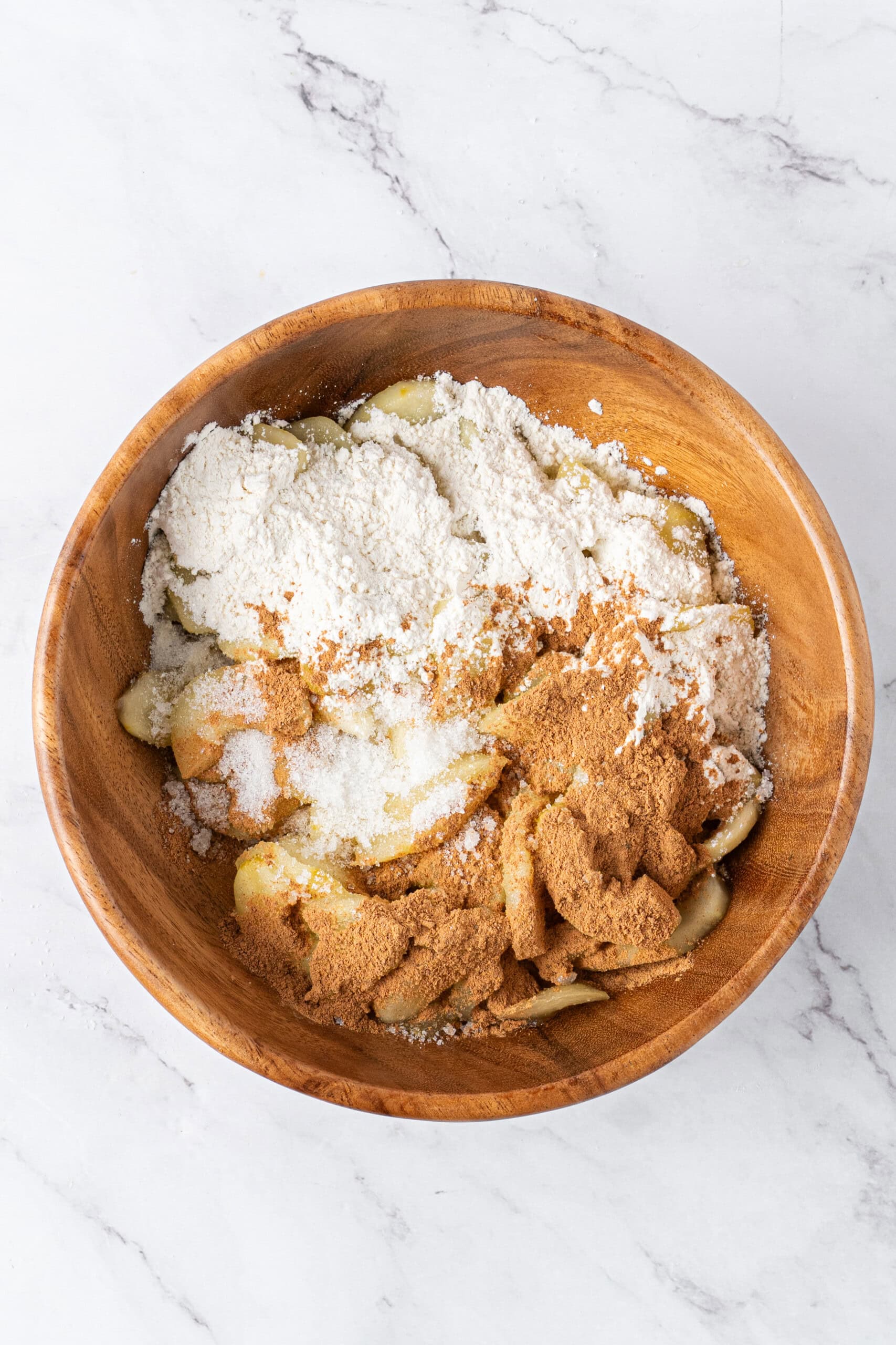 Cooked apple slices topped with flour, cinnamon, and sugar in a wooden mixing bowl.