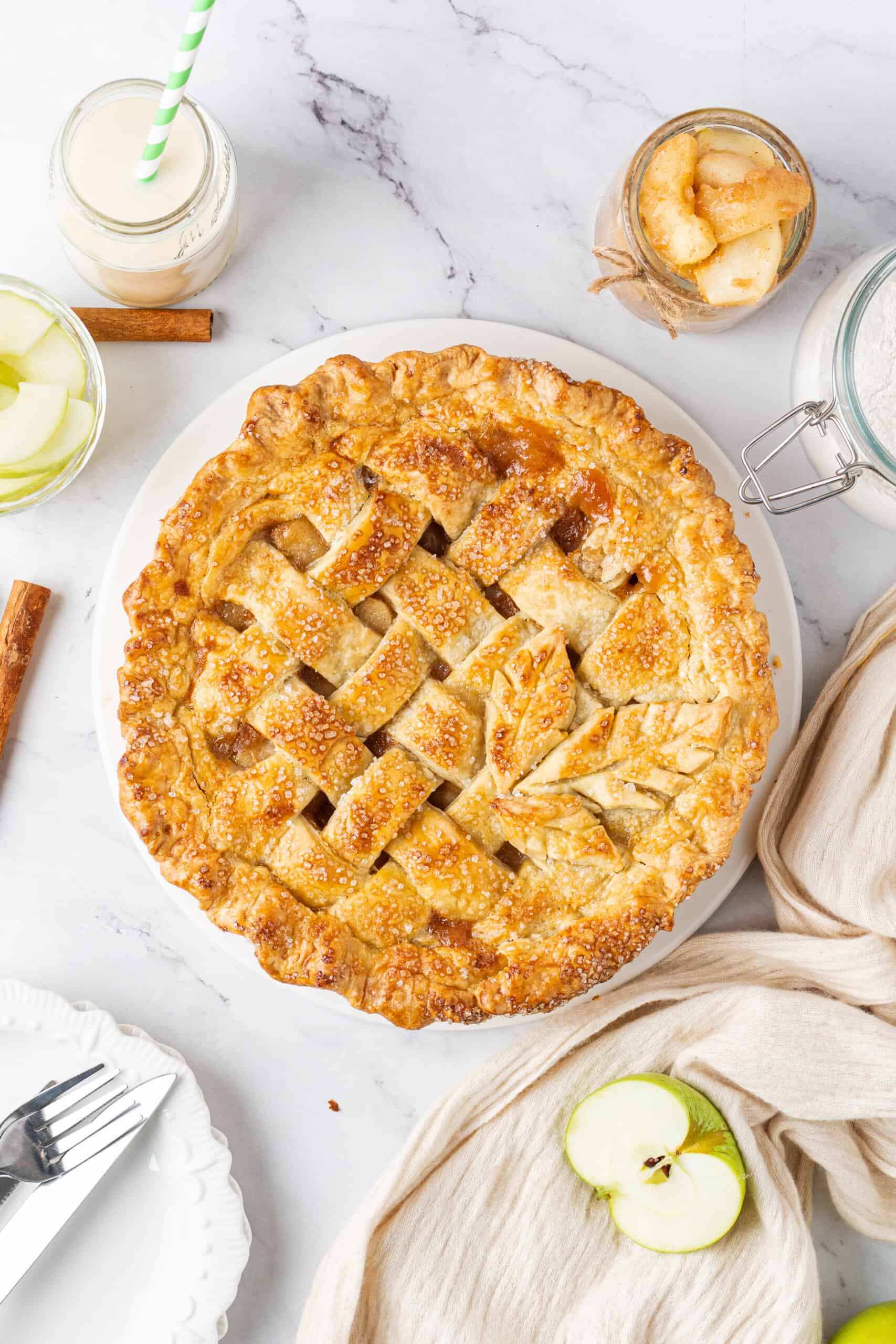 User attachment Homemade apple pie with a golden brown lattice crust, coarse sugar topping, and decorative pastry leaves, surrounded by fresh apples and cinnamon sticks.