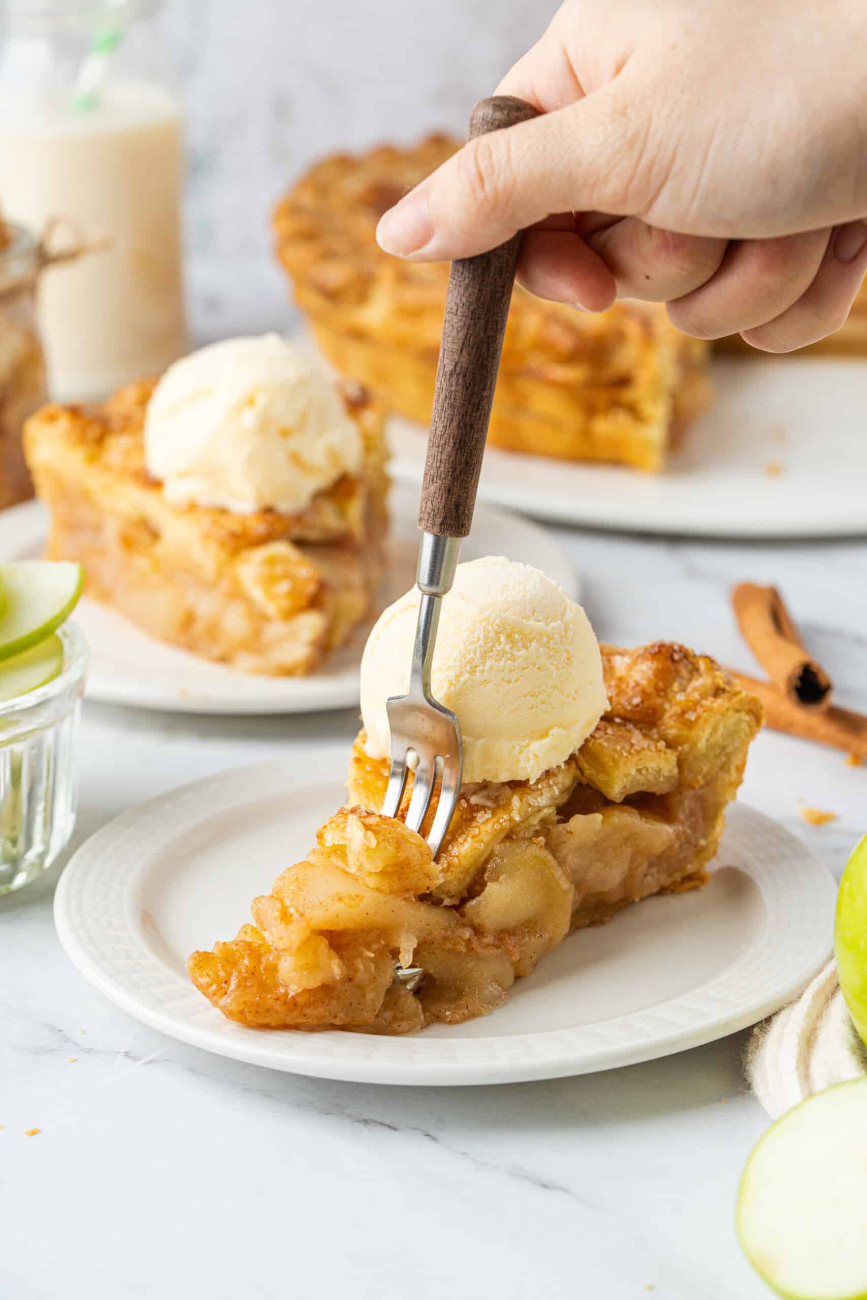 A slice of homemade apple pie with tender, cinnamon-spiced apple filling and a golden, flaky crust, topped with vanilla ice cream as a fork takes a bite.