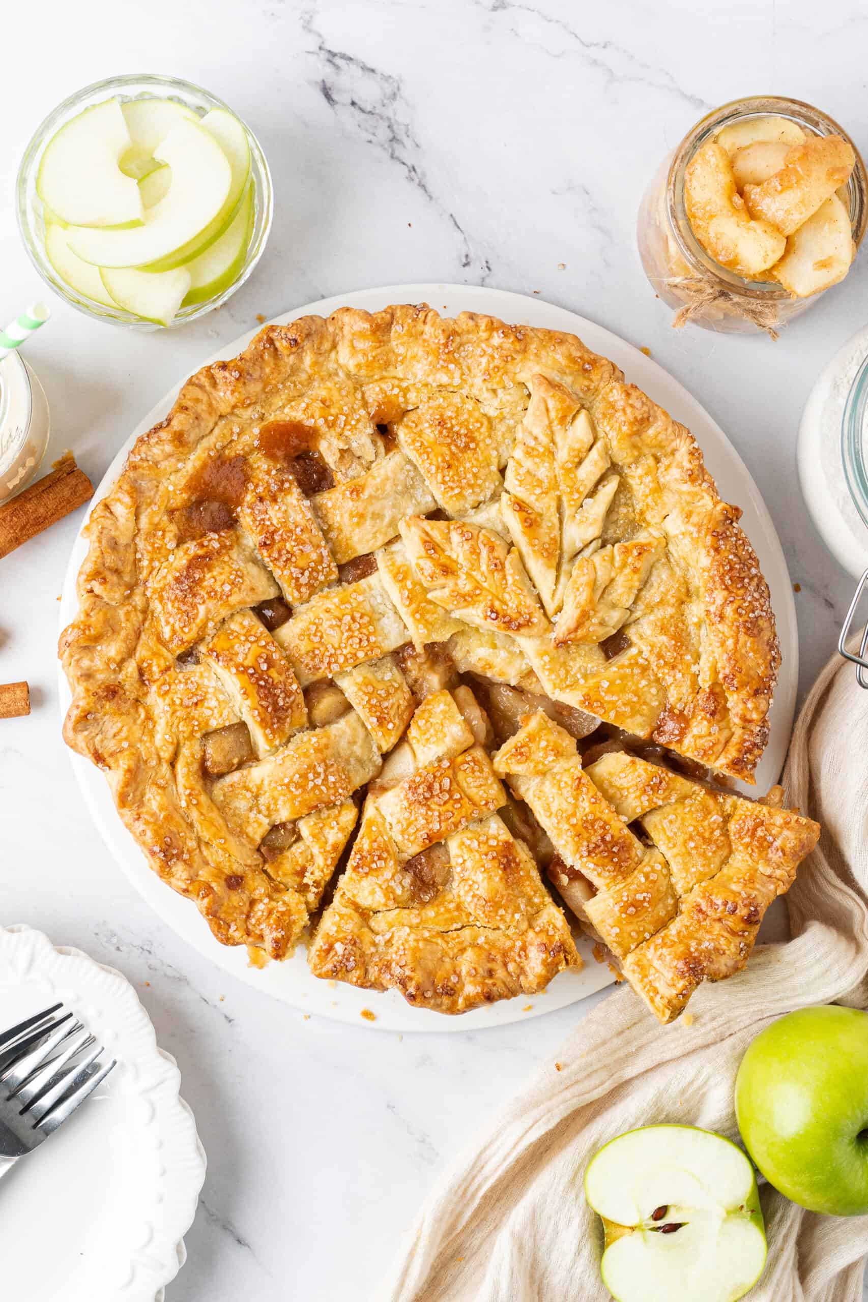 Overhead view of an apple pie with a golden, flaky lattice crust sprinkled with coarse sugar, showing slices cut to reveal tender cinnamon-spiced apple filling, surrounded by fresh green apples and cinnamon sticks.
