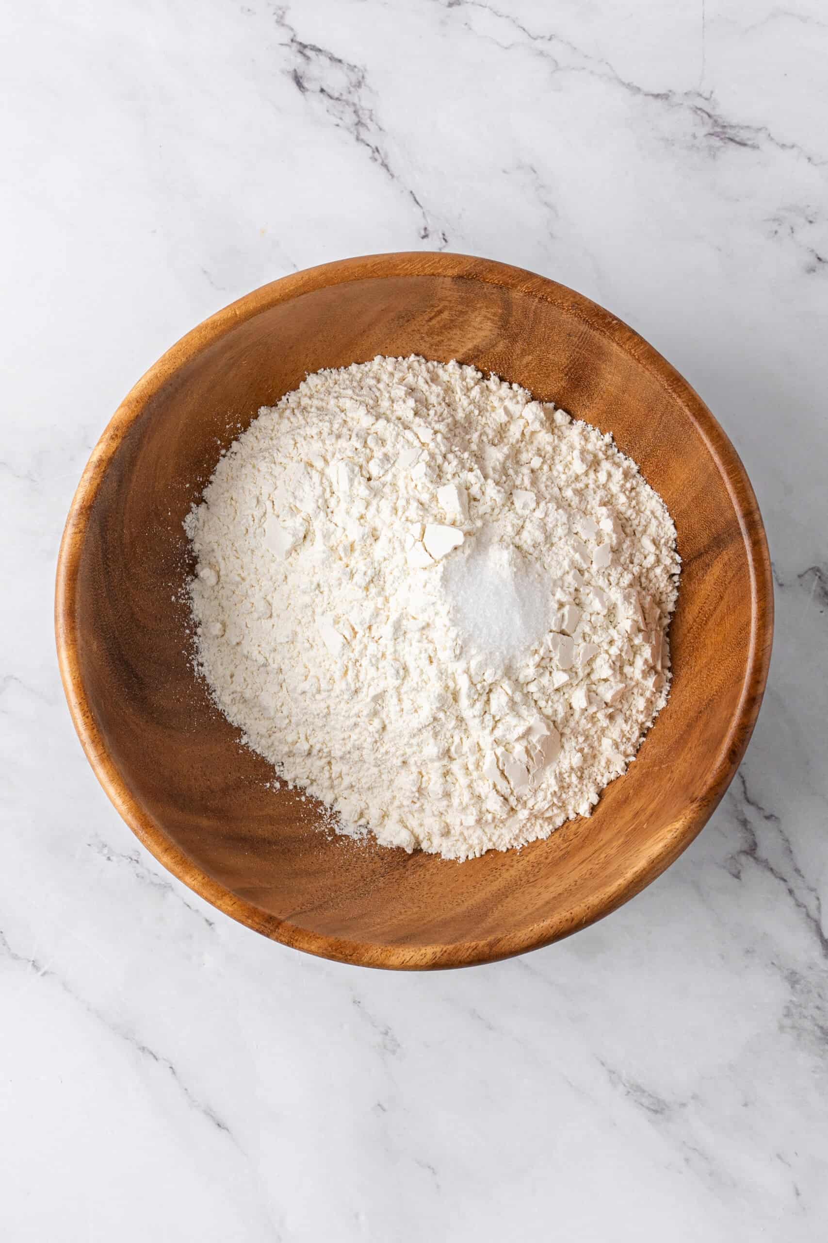 A wooden bowl filled with flour and dry ingredients on a white marble countertop.