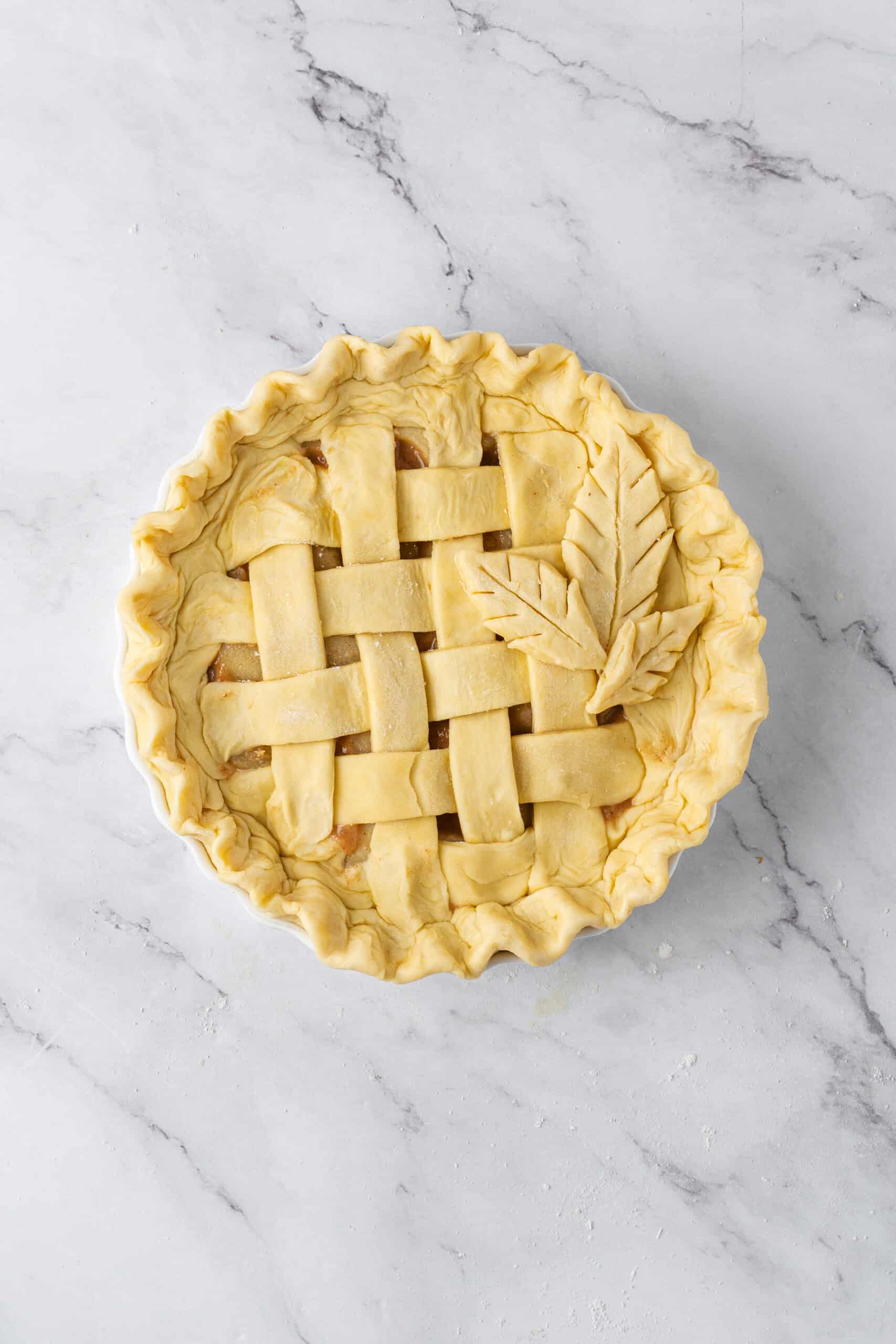 Unbaked pie with a woven lattice crust, decorative pastry leaves, and crimped edges in a white pie dish.